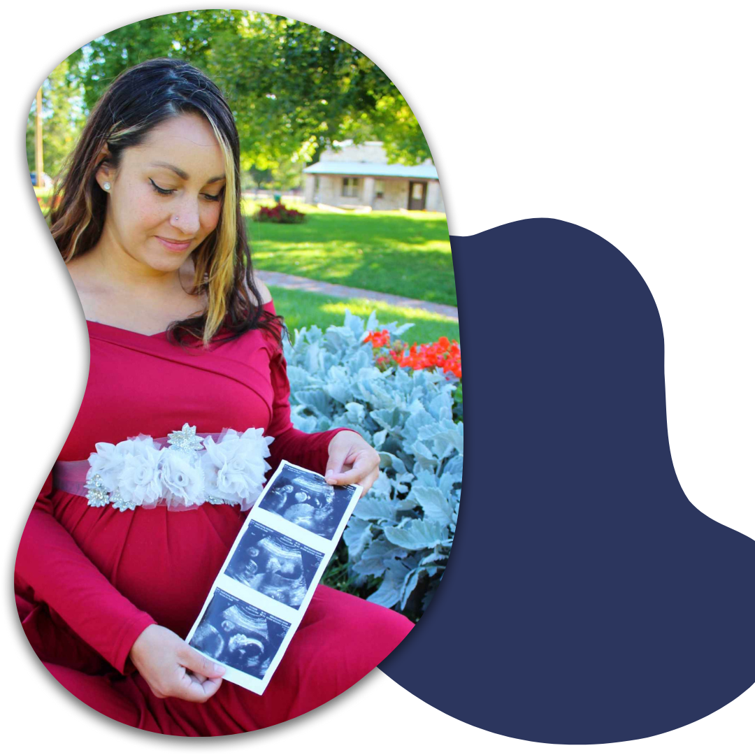 Pregnant woman in a red dress sitting outdoors holding ultrasound images, with greenery and a house in the background.