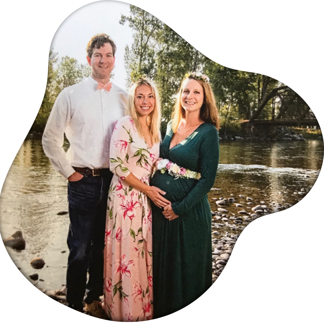 Three people, a man and two women, standing by a river, smiling. The pregnant woman is wearing a green dress with a floral belt, and the other woman is in a pink floral dress. The man is in a white shirt with a peach-colored bow tie. Trees and a bridge are in the background in a natural outdoor setting.