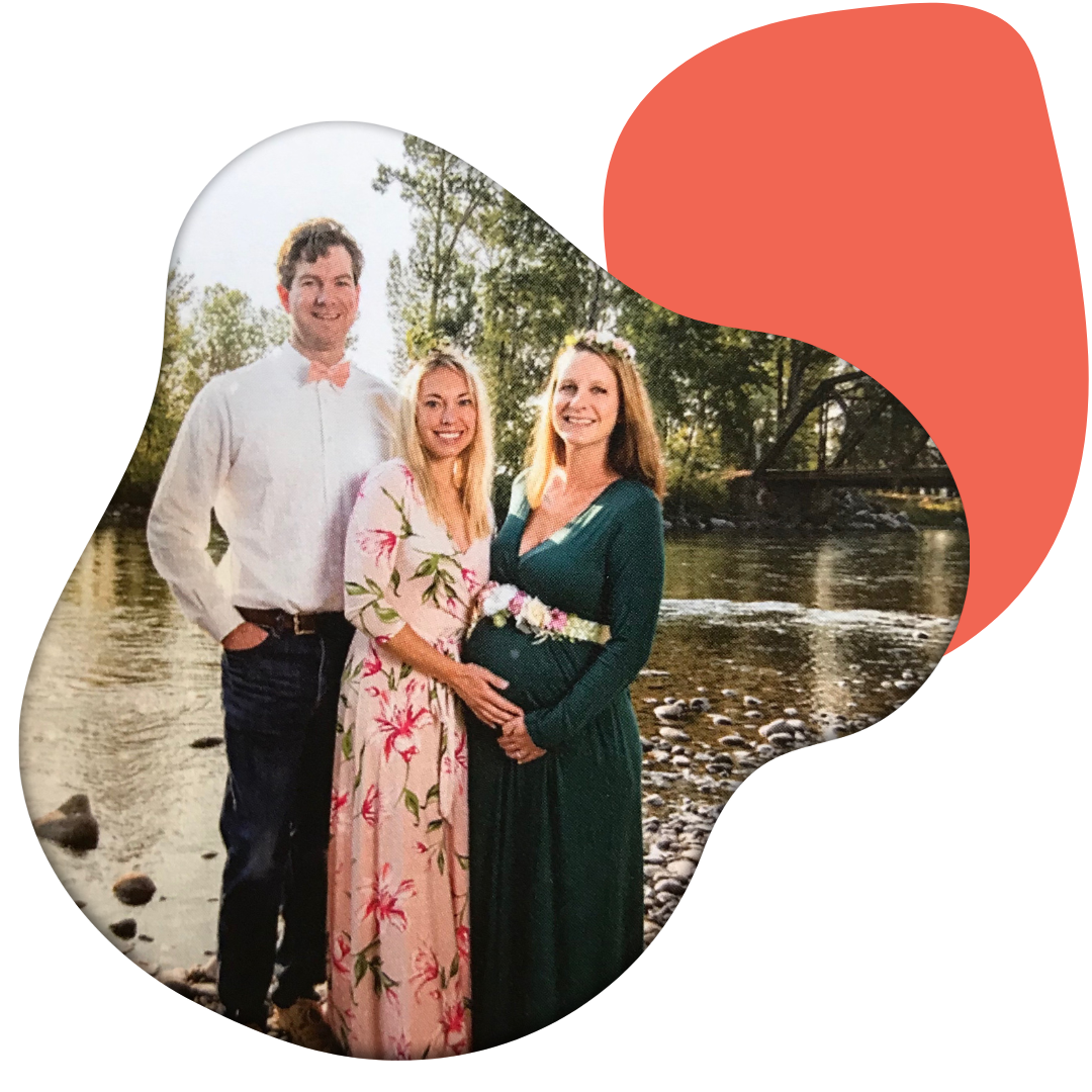 Three people, a man and two women, standing by a river with trees in the background, smiling at the camera. One woman is pregnant, wearing a floral dress; the other woman is in a dark green dress with a floral belt; the man is in a white shirt and dark pants.