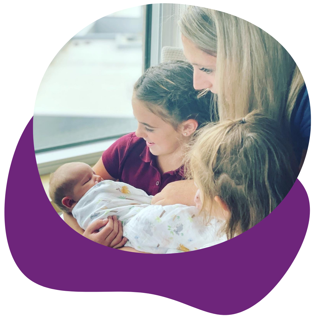 A woman holding a newborn baby with two other women touching the baby, near a window in a hospital or medical setting.