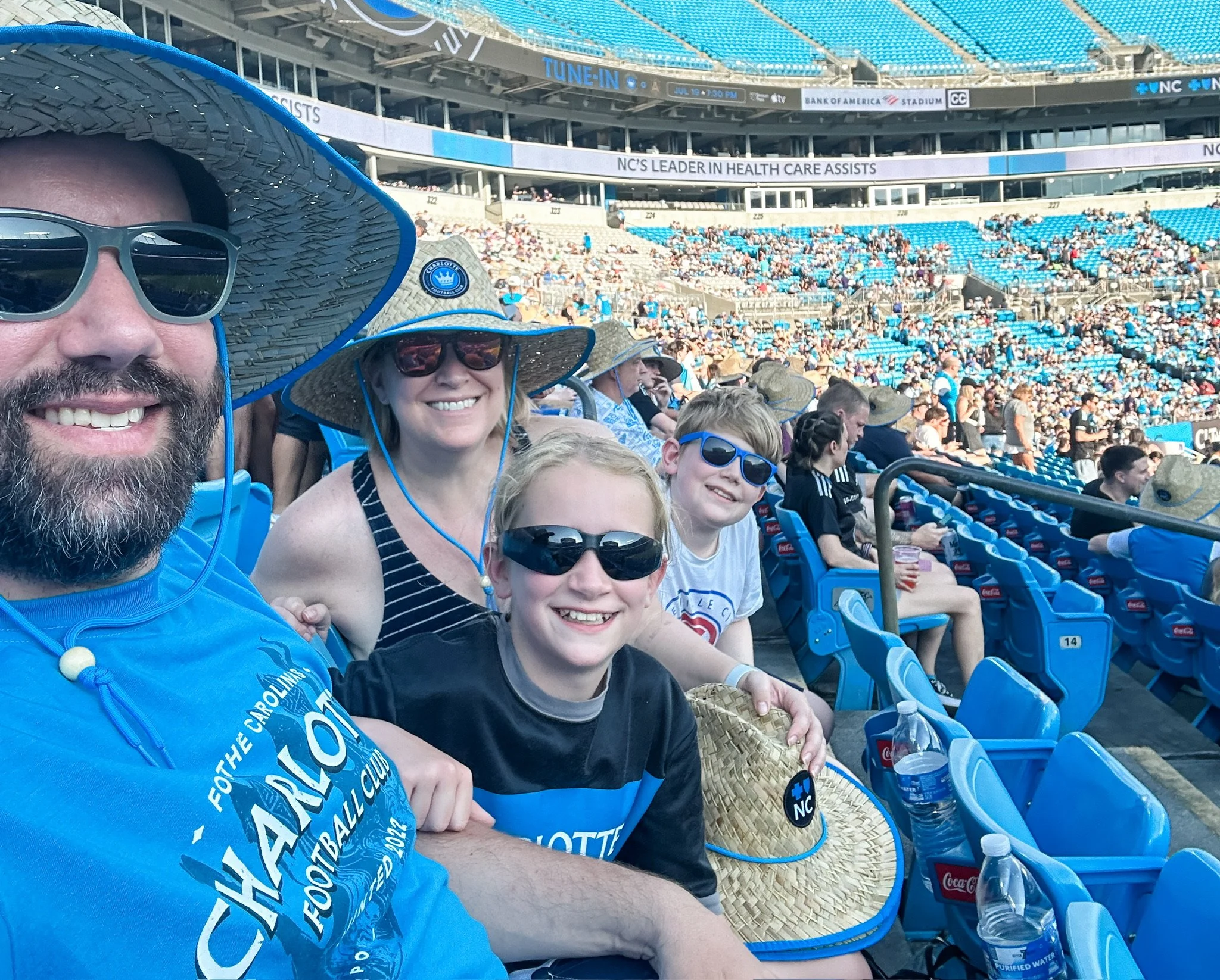 A group of people, including two adults and two children, sitting at a stadium with blue seats filling most of the background. The people are wearing sun hats and sunglasses, and there are water bottles on the seat in front of them. The stadium has a