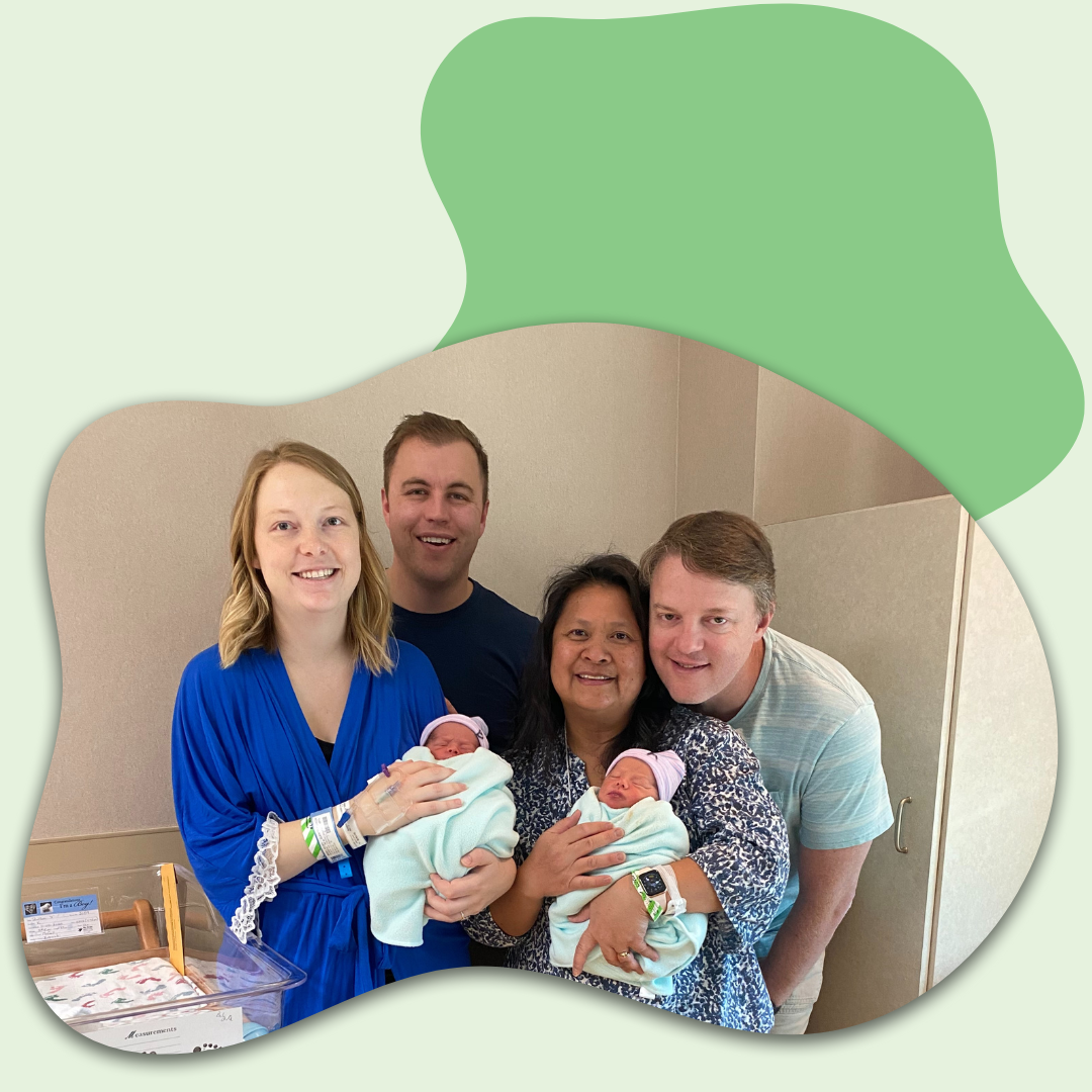Two women holding newborn babies standing with two men, all smiling indoors in a hospital or birthing center room.