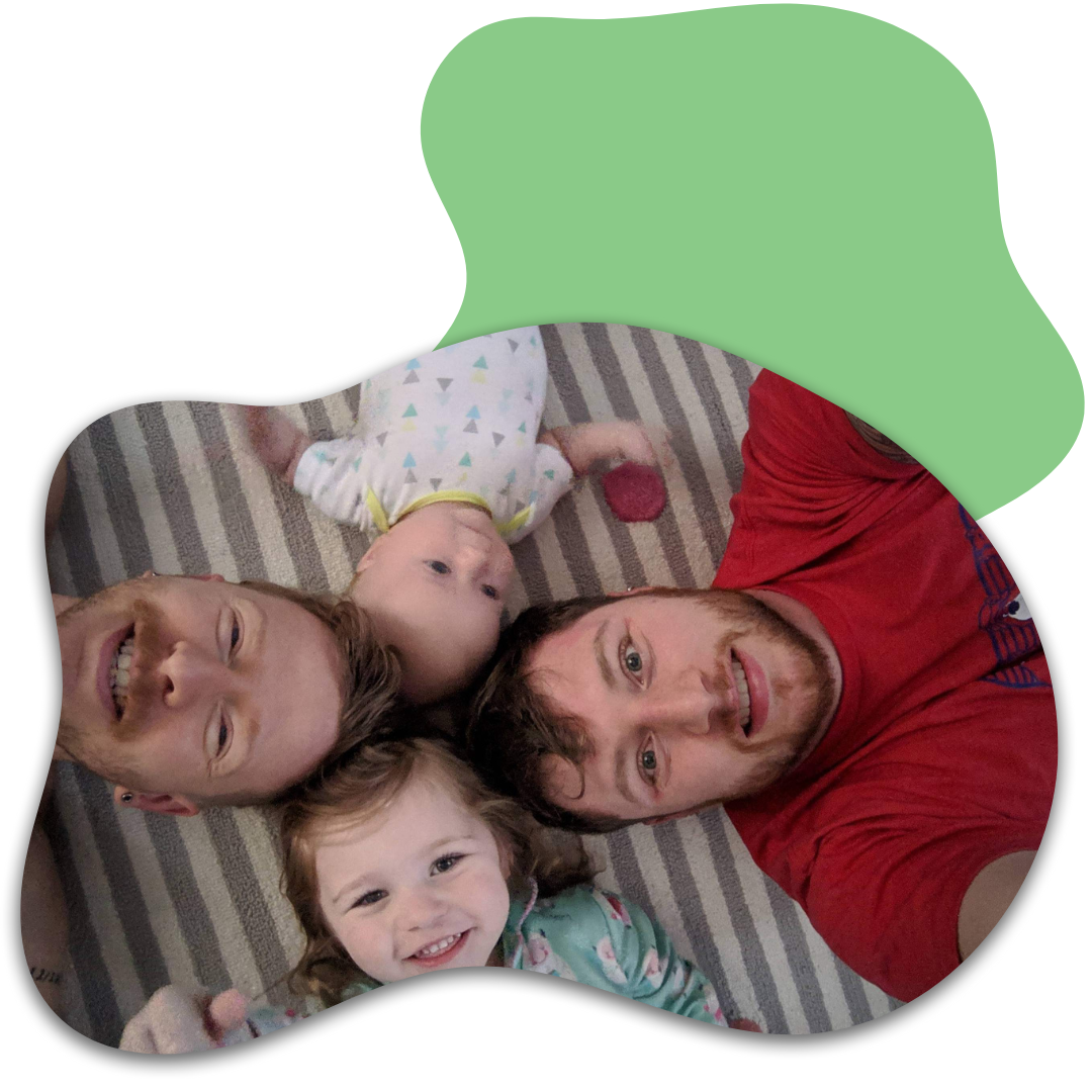 A man and two young children lying on a striped carpet, looking up and smiling at the camera. The man has dark hair and a red shirt, the girl has light brown hair and is smiling, and the baby has light-colored hair and is wearing a light-colored onesie with a pattern.