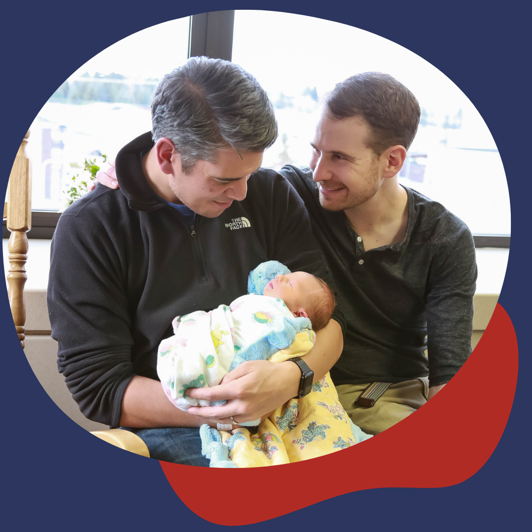 Two men hold a newborn baby in a hospital room, smiling and looking at each other.