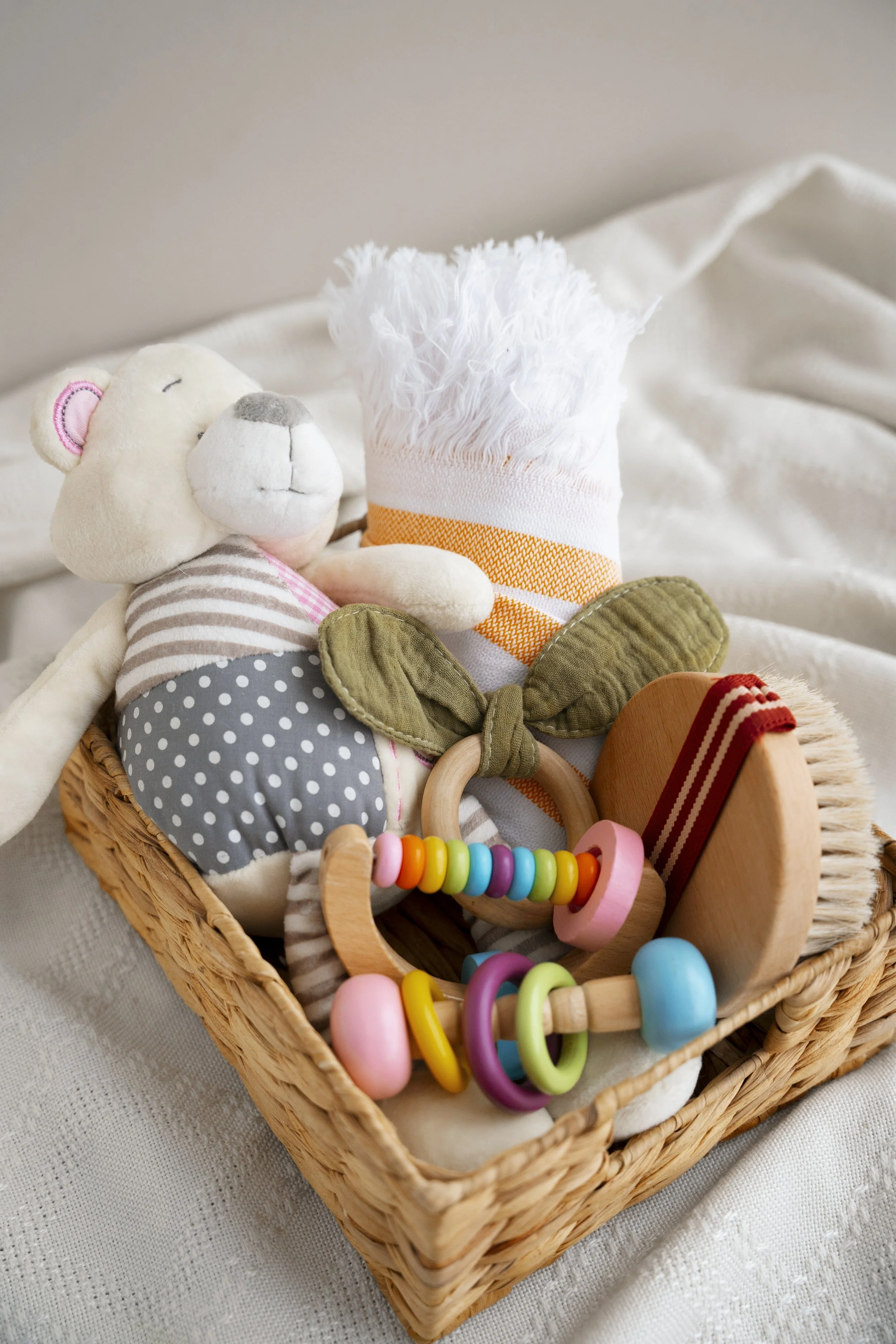 A wicker basket filled with soft toys and baby's teething toys, including a plush bear, a white sock with orange stripes, and colorful silicone rings, resting on a white fabric surface.