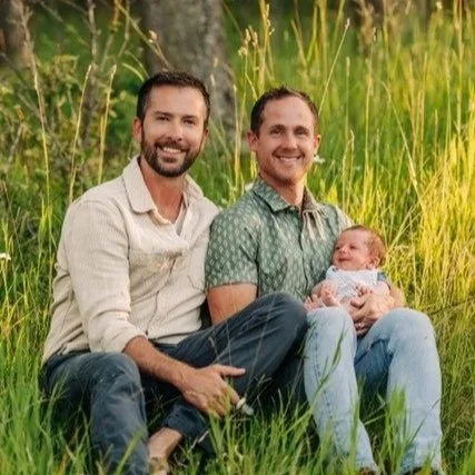 Two men and a baby sitting in tall grass outdoors with trees in the background.