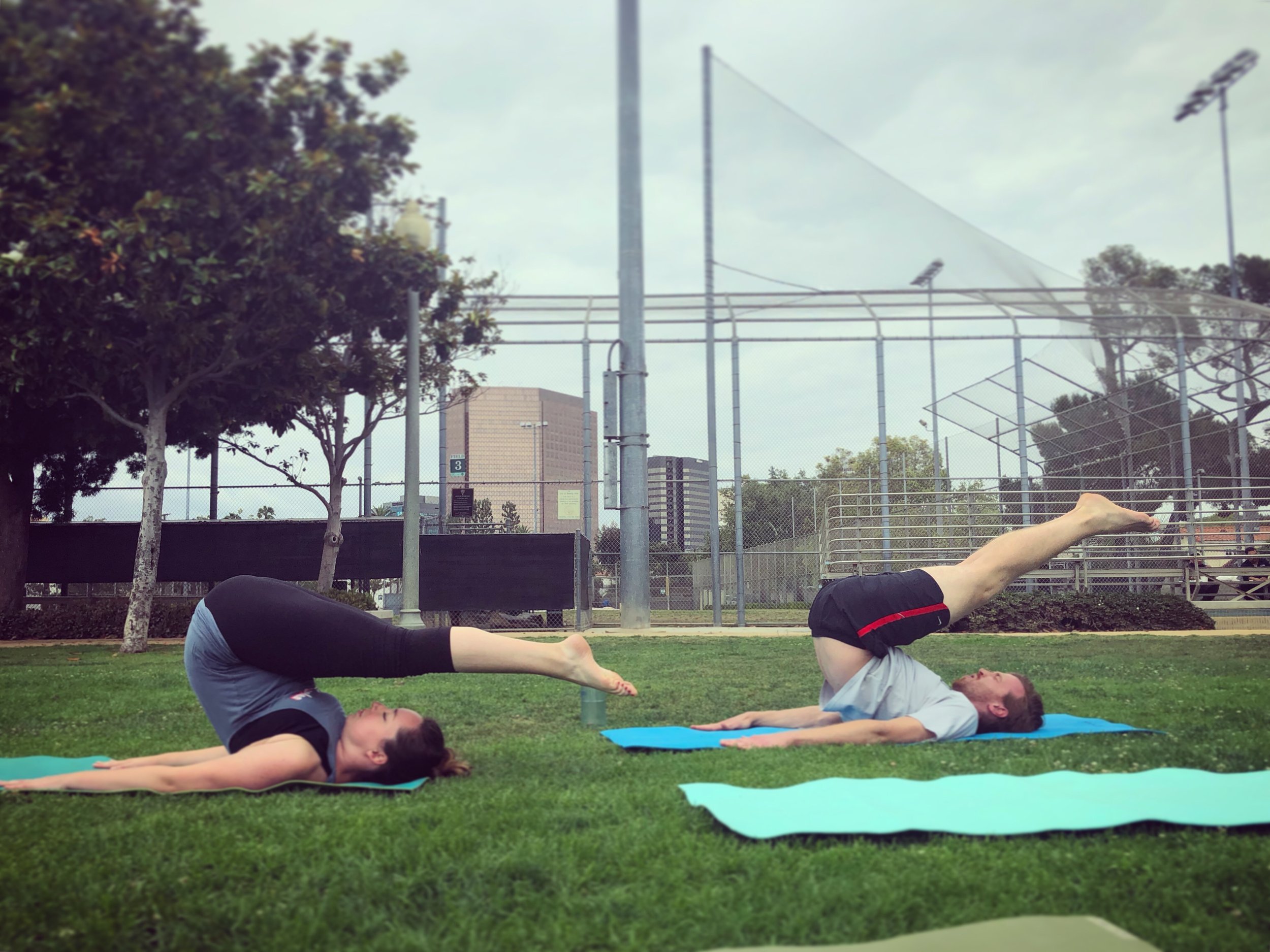 A group of people participating in a community Pilates class in a warmly lit studio, performing stretching poses supported by a foam roller, lying on their backs on Pilates mats.