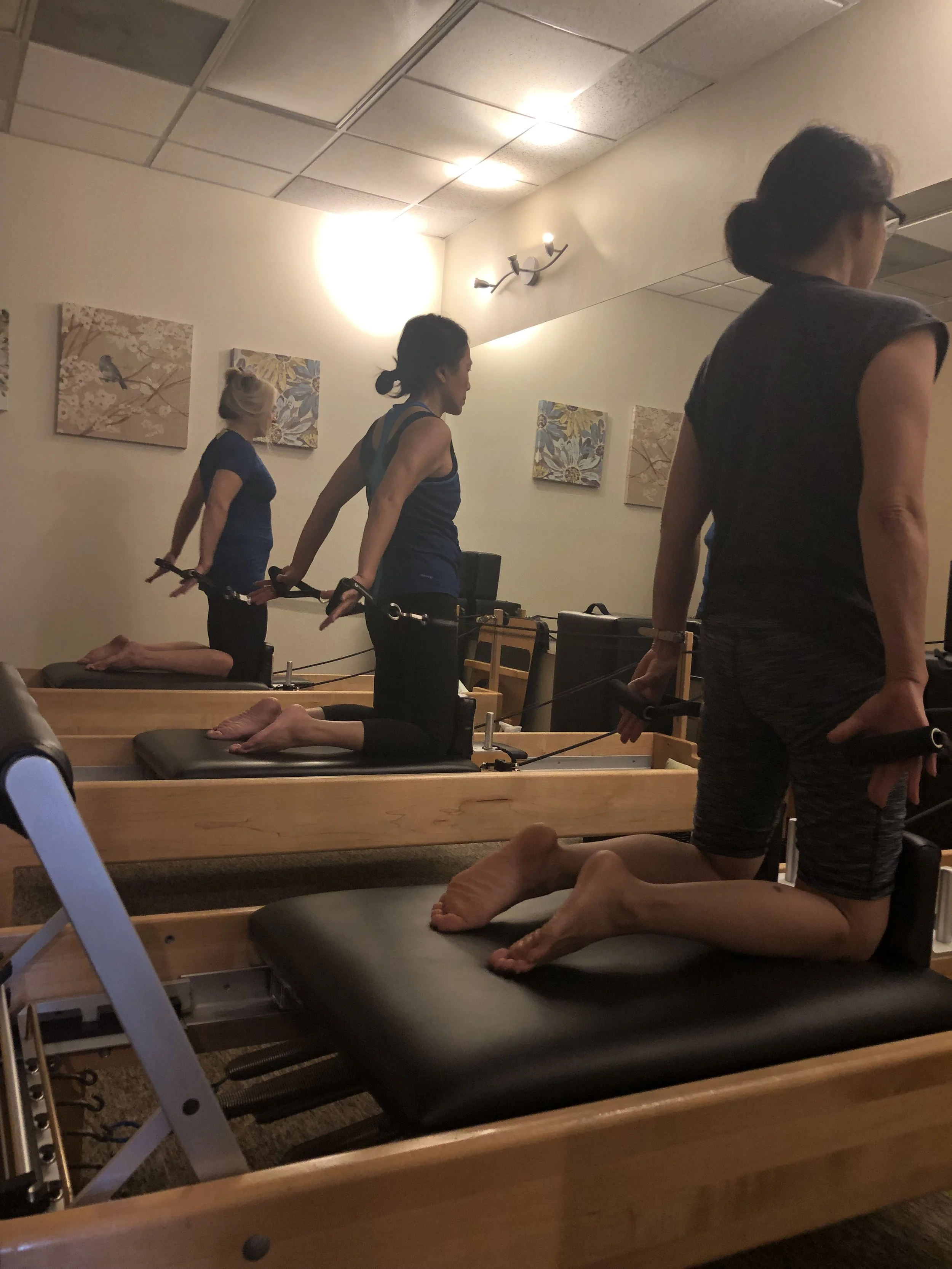 Three women using Pilates reformer machines in a fitness studio, with two women kneeling on the reformers and one woman standing, all holding handles and engaging in a Pilates exercise.