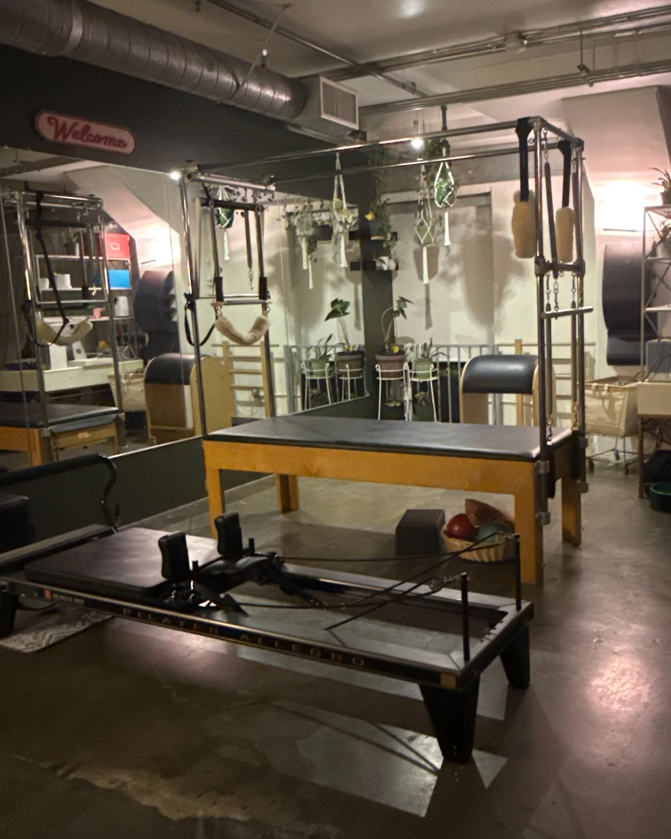Indoor Pilates studio with Pilates reformer machine, pilates tower, and other fitness equipment, decorated with hanging plants and a welcome sign.