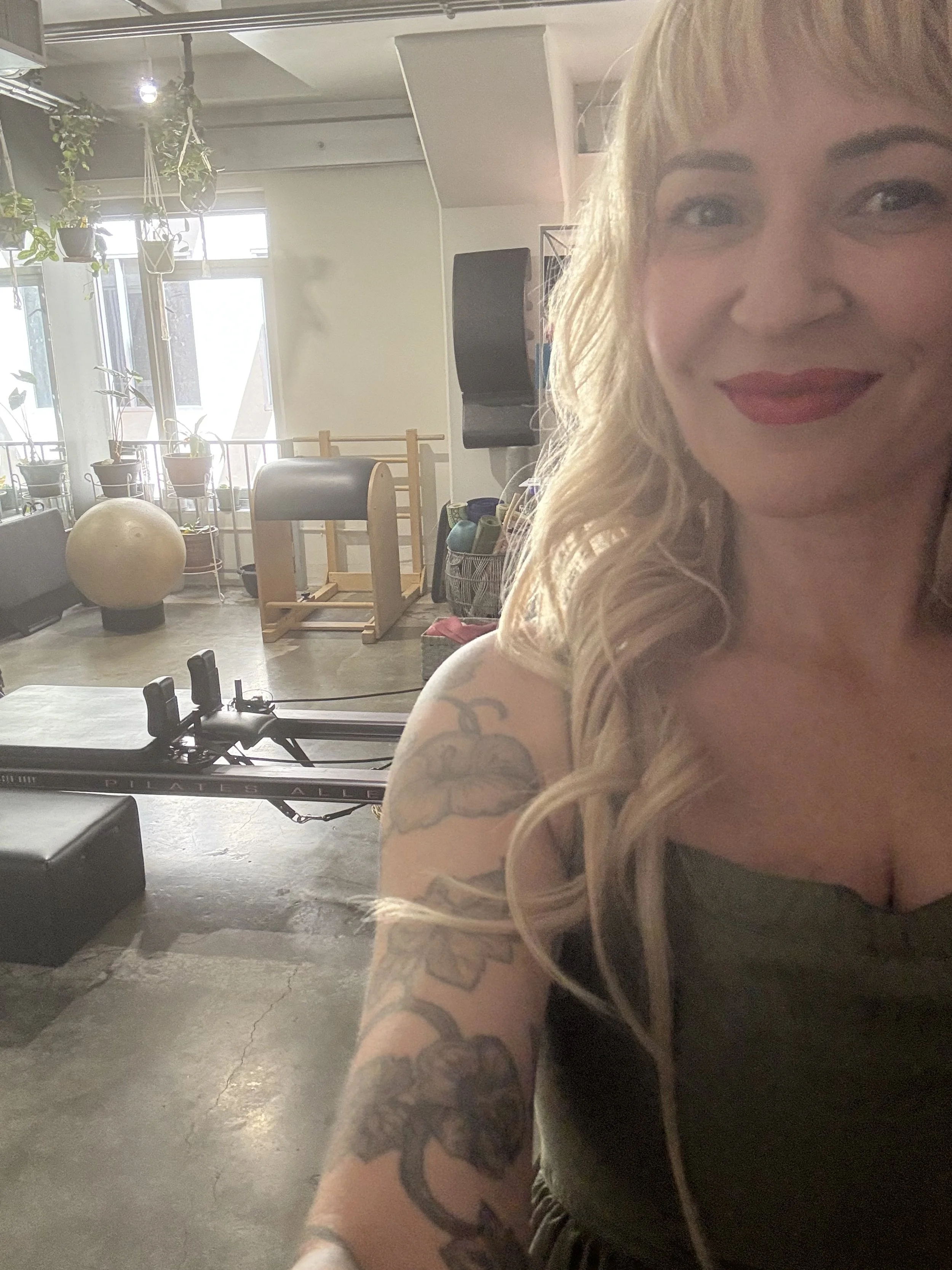 Close-up of a smiling Pilates Instrctor in a private studio with Pilates equipment, plants, and sunlight in the background.