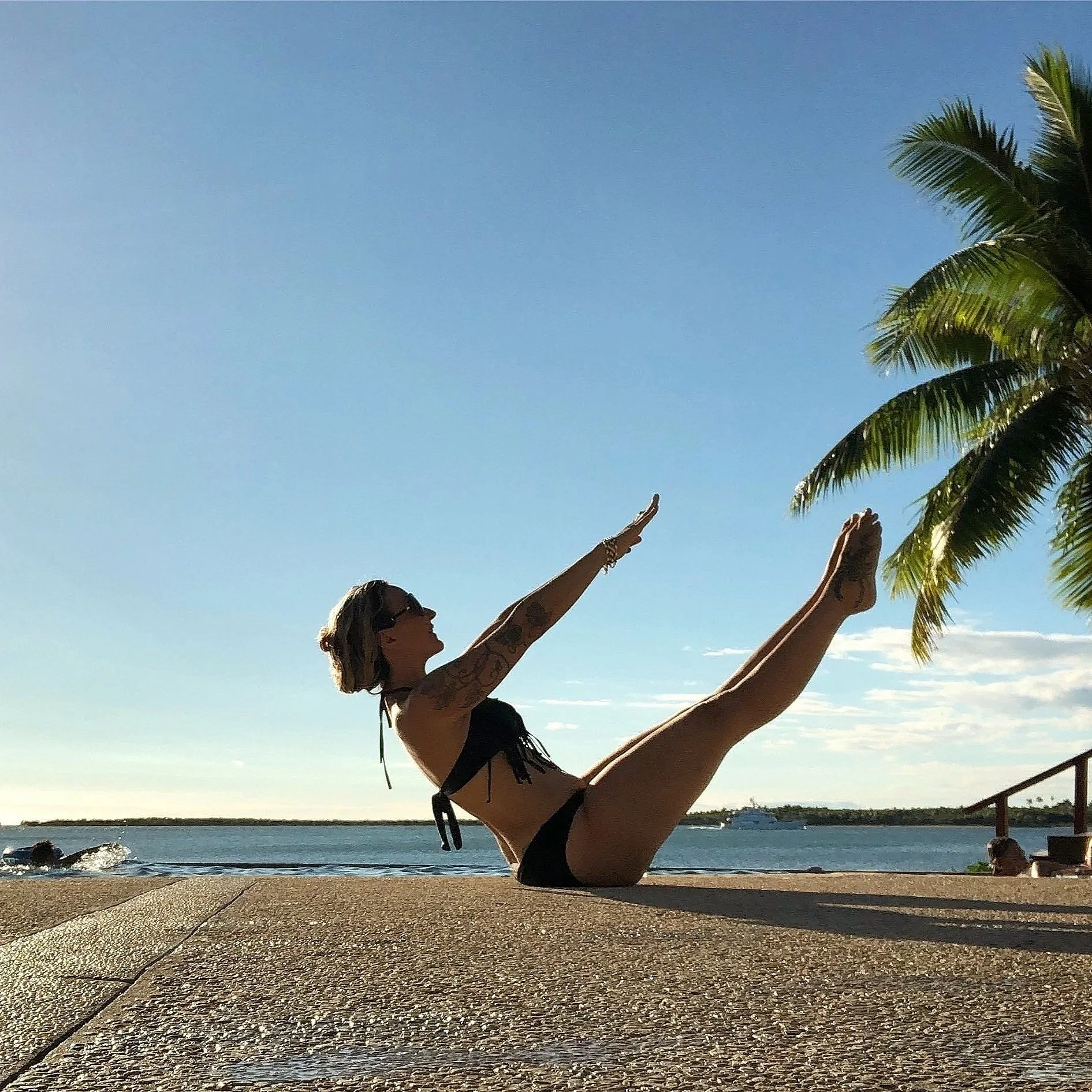 A woman in a black bikini doing Pilates outdoors by the water, with palm trees and a boat in the background.
