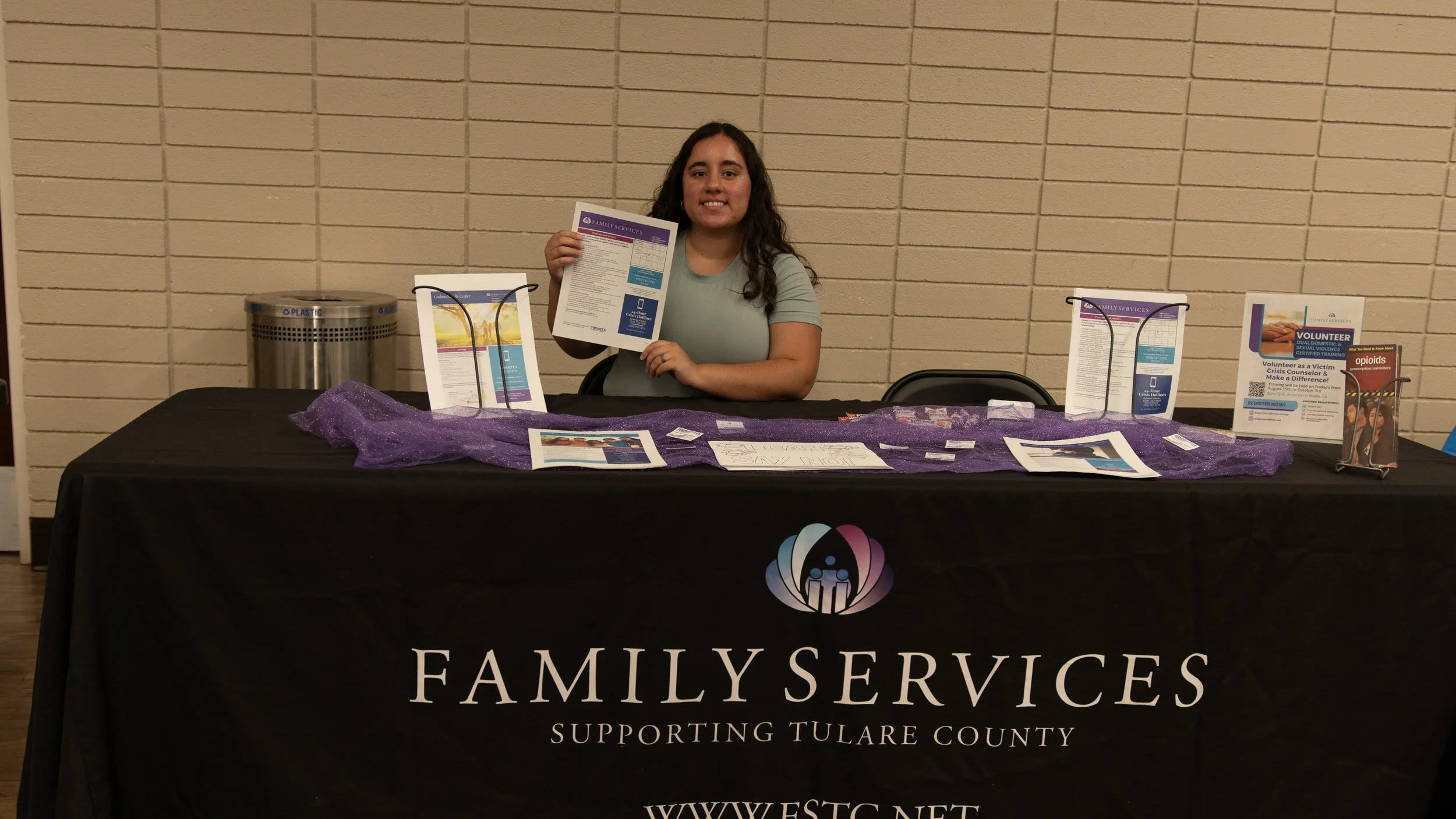 A woman sitting at a table with informational pamphlets and materials related to Family Services, supporting Tulare County, with a purple cloth and signs on the table.