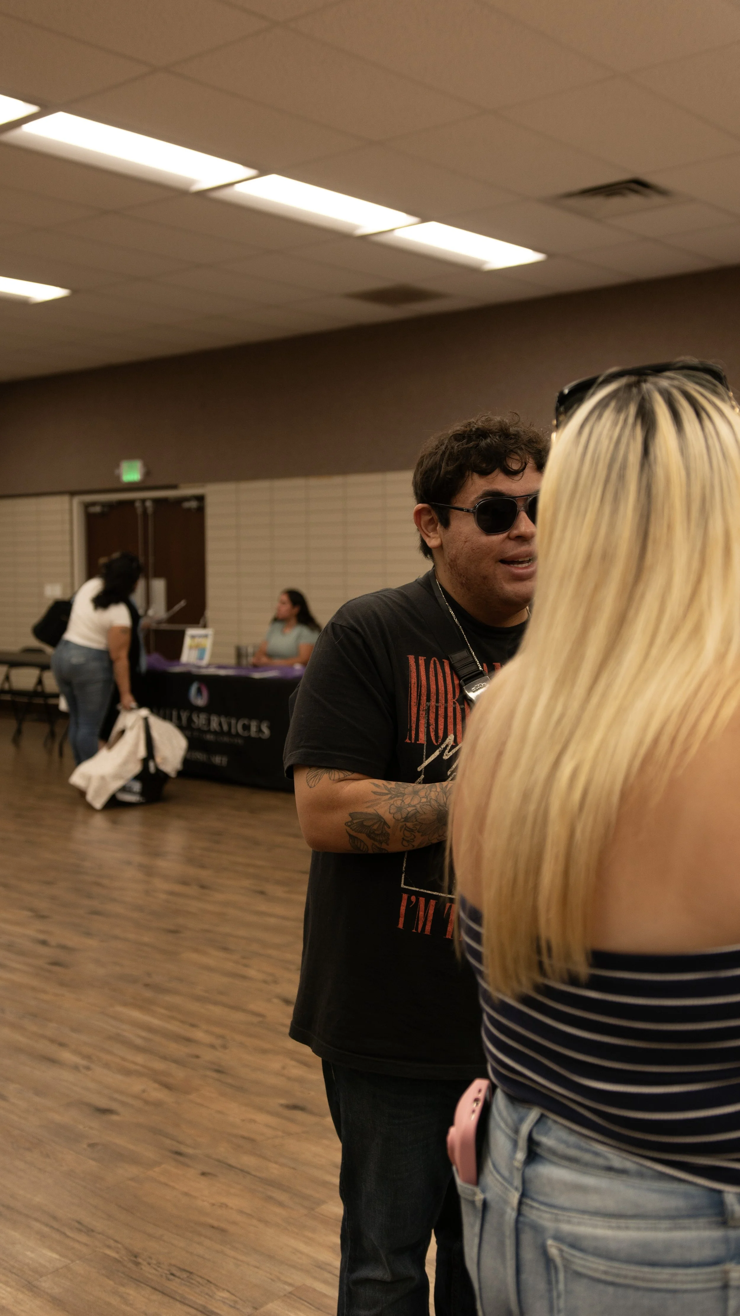 Two people having a conversation in a room with a wooden floor and a drop ceiling. One person with dark hair, sunglasses, and tattoos is speaking to a blonde woman in a striped shirt whose back is to the camera. In the background, a woman is standing
