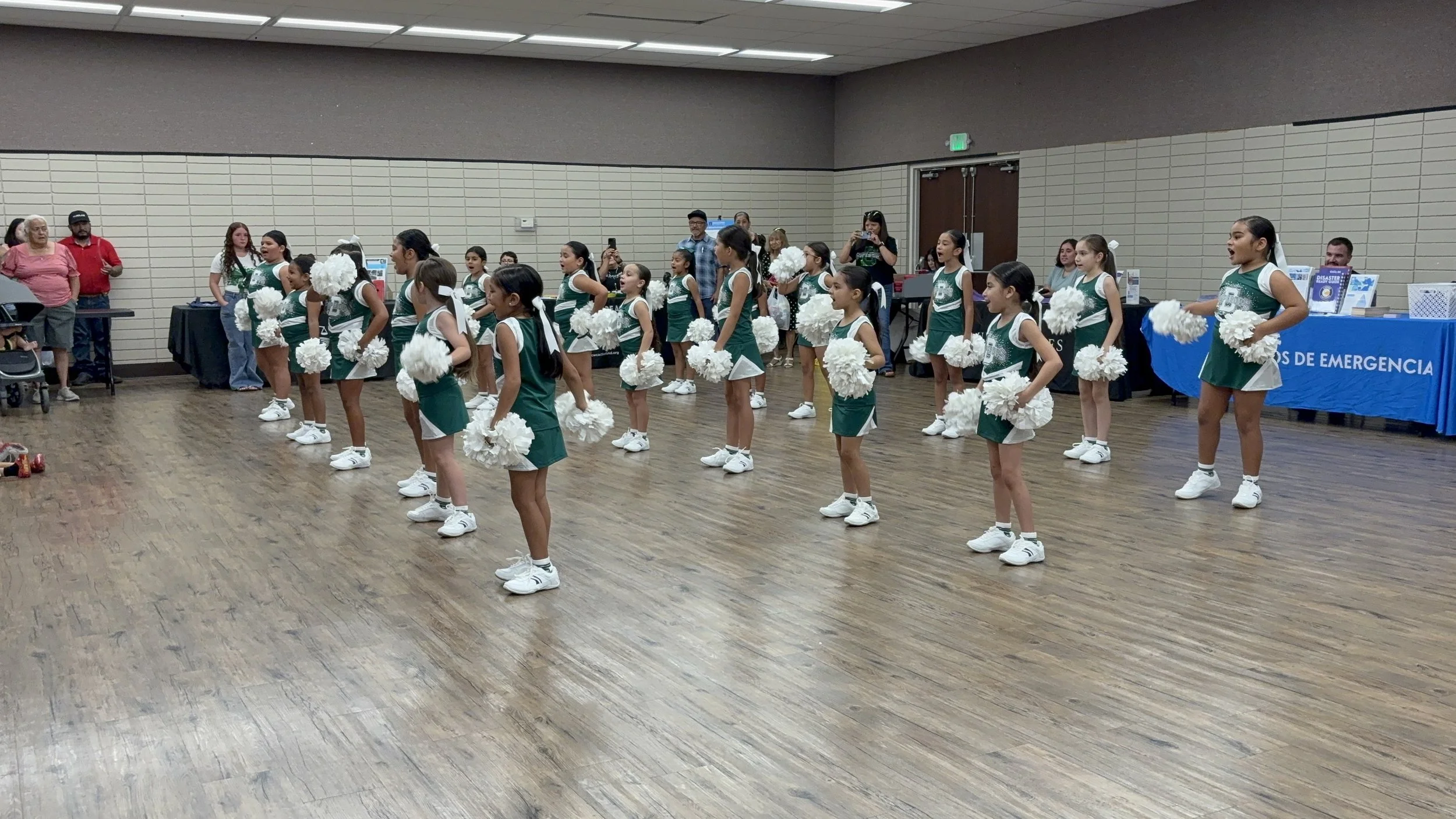 Young cheerleaders in green and white uniforms performing with pom-poms in a room with wood floors and beige walls, audience behind them taking photos.
