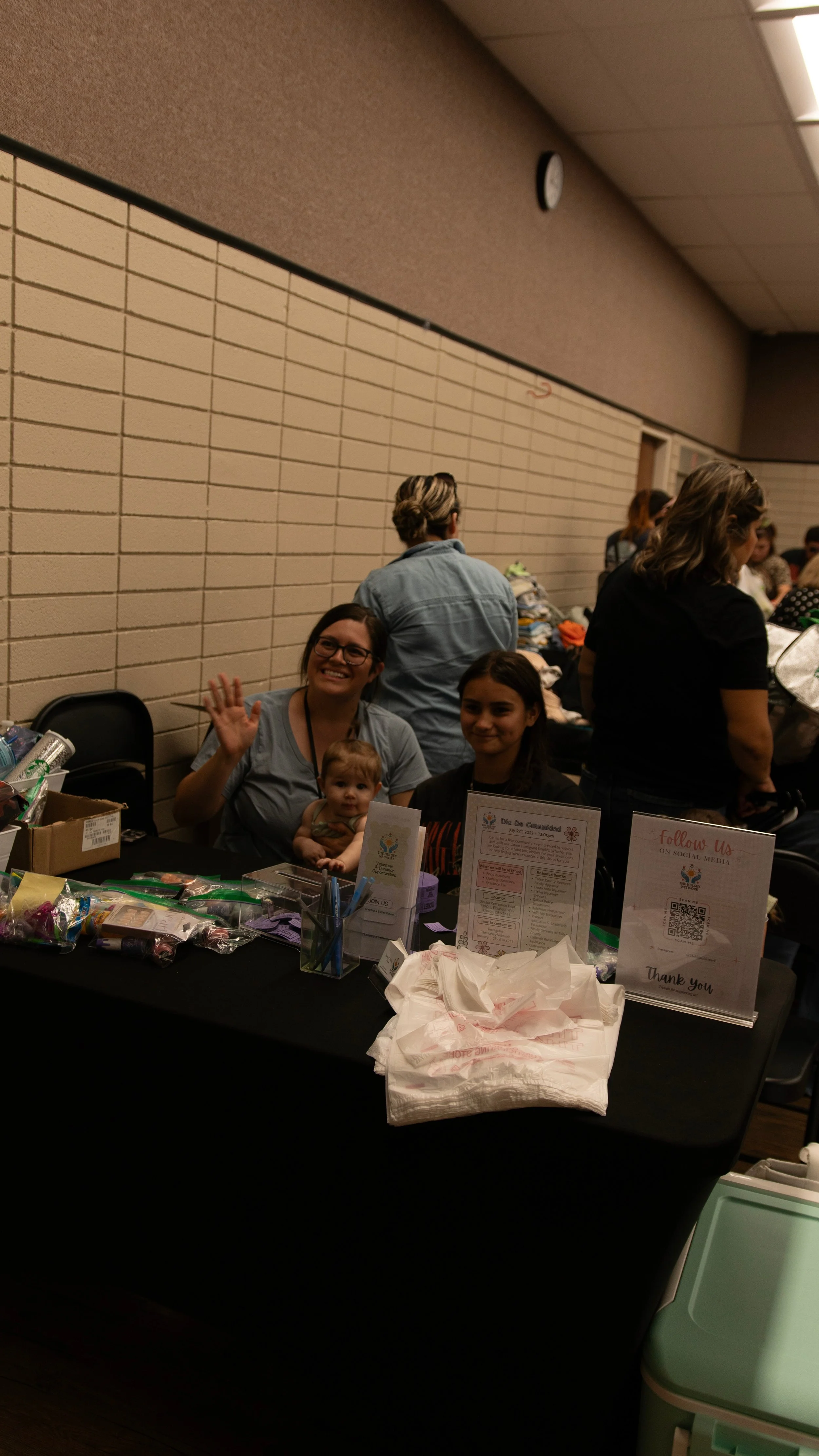 A group of people at a community event booths, including a woman with glasses and a baby, sitting at a table with informational signs, supplies, and a thank you sign.