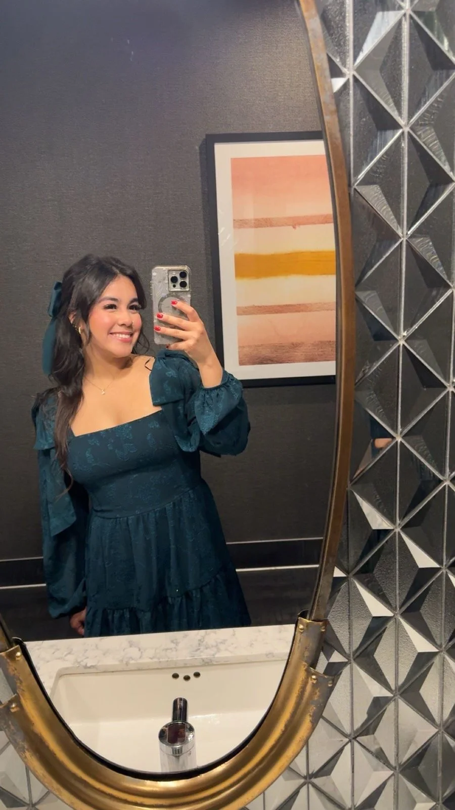 A woman taking a selfie in a bathroom mirror. She has dark wavy hair, wears a teal dress with puffed sleeves, and is smiling. The bathroom has a textured dark gray wall, a framed abstract art piece with horizontal bands of pink, yellow, and brown, and a geometric decorative mirror frame with triangle and diamond shapes.
