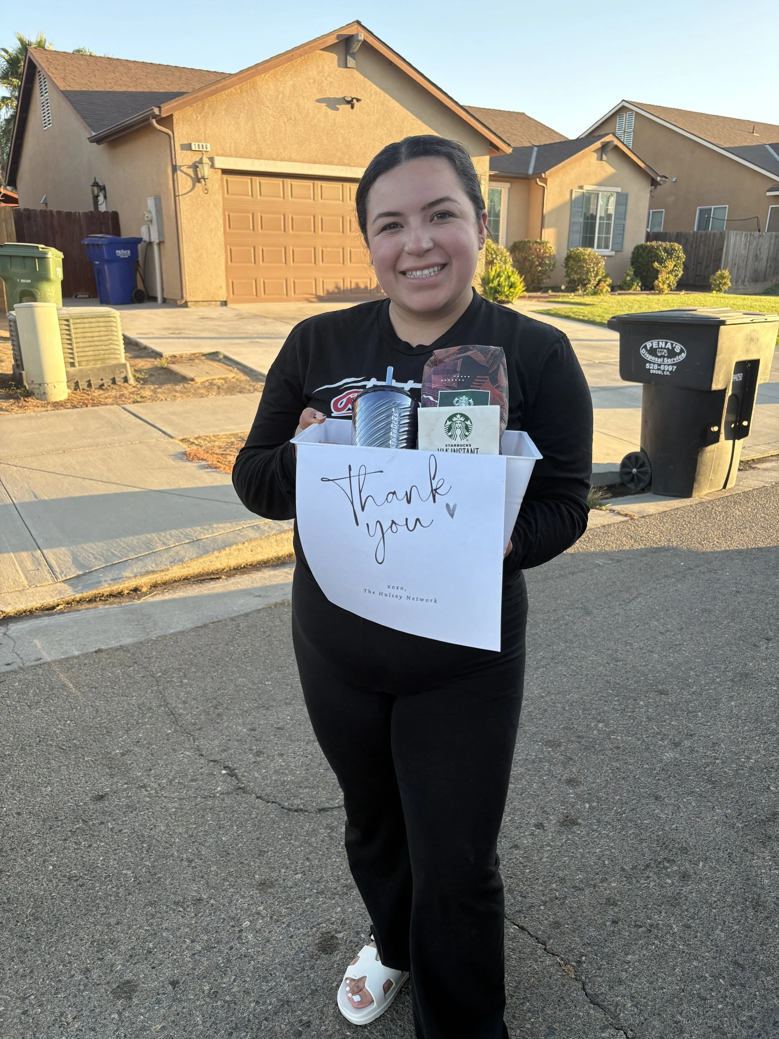 A young woman standing on a suburban street holding a gift basket with a Starbucks cup, a thank you note, and some items inside. She's smiling and wearing a black shirt and black pants, with houses and trash bins visible in the background.
