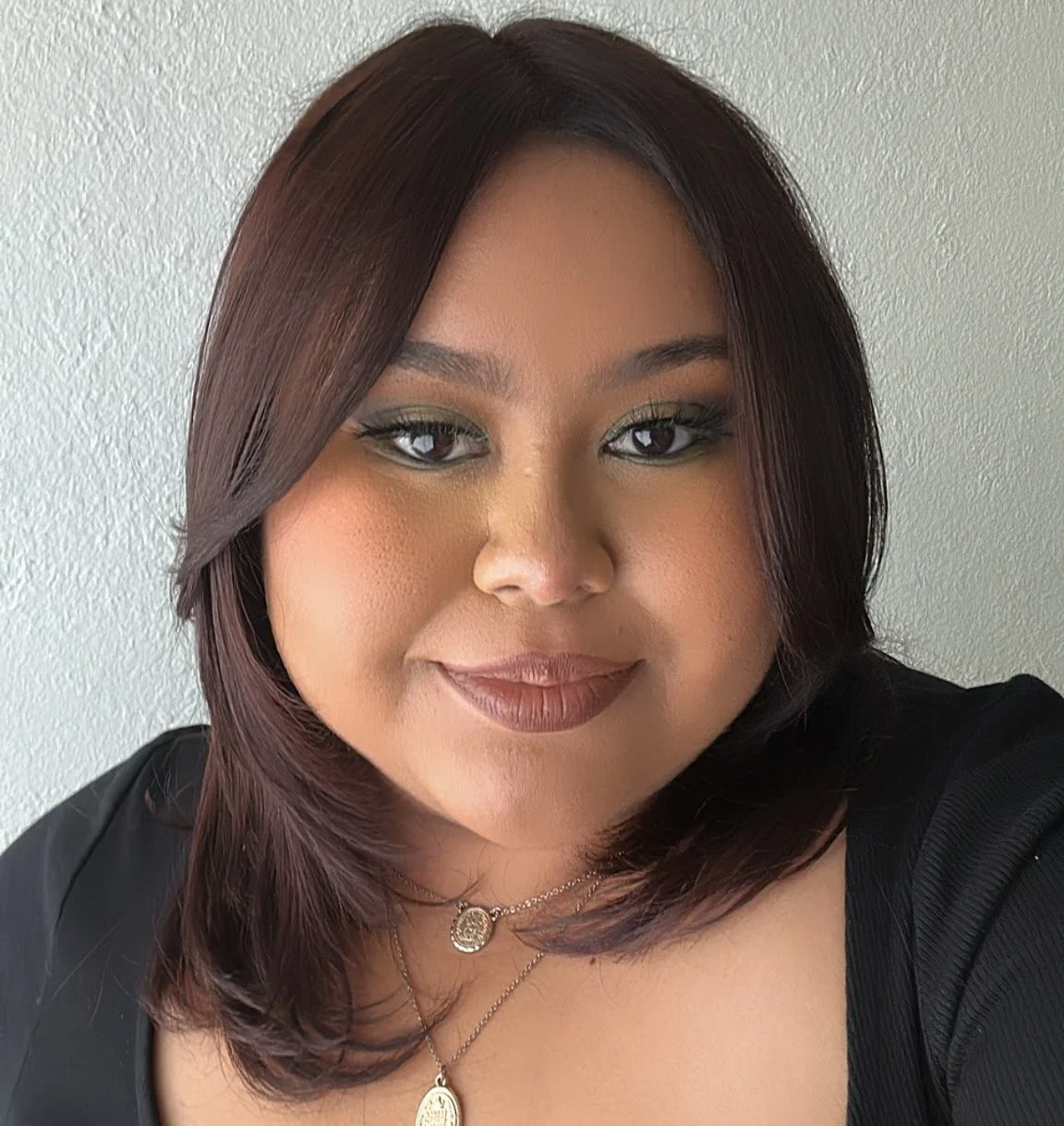 Close-up of a woman with shoulder-length dark hair, makeup, and wearing layered necklaces, smiling slightly in front of a plain white wall.