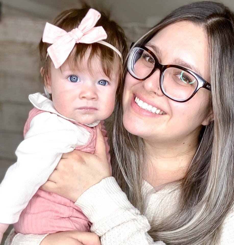 A woman with glasses smiling and holding a baby girl with blue eyes, a pink bow, and a pink and white outfit.