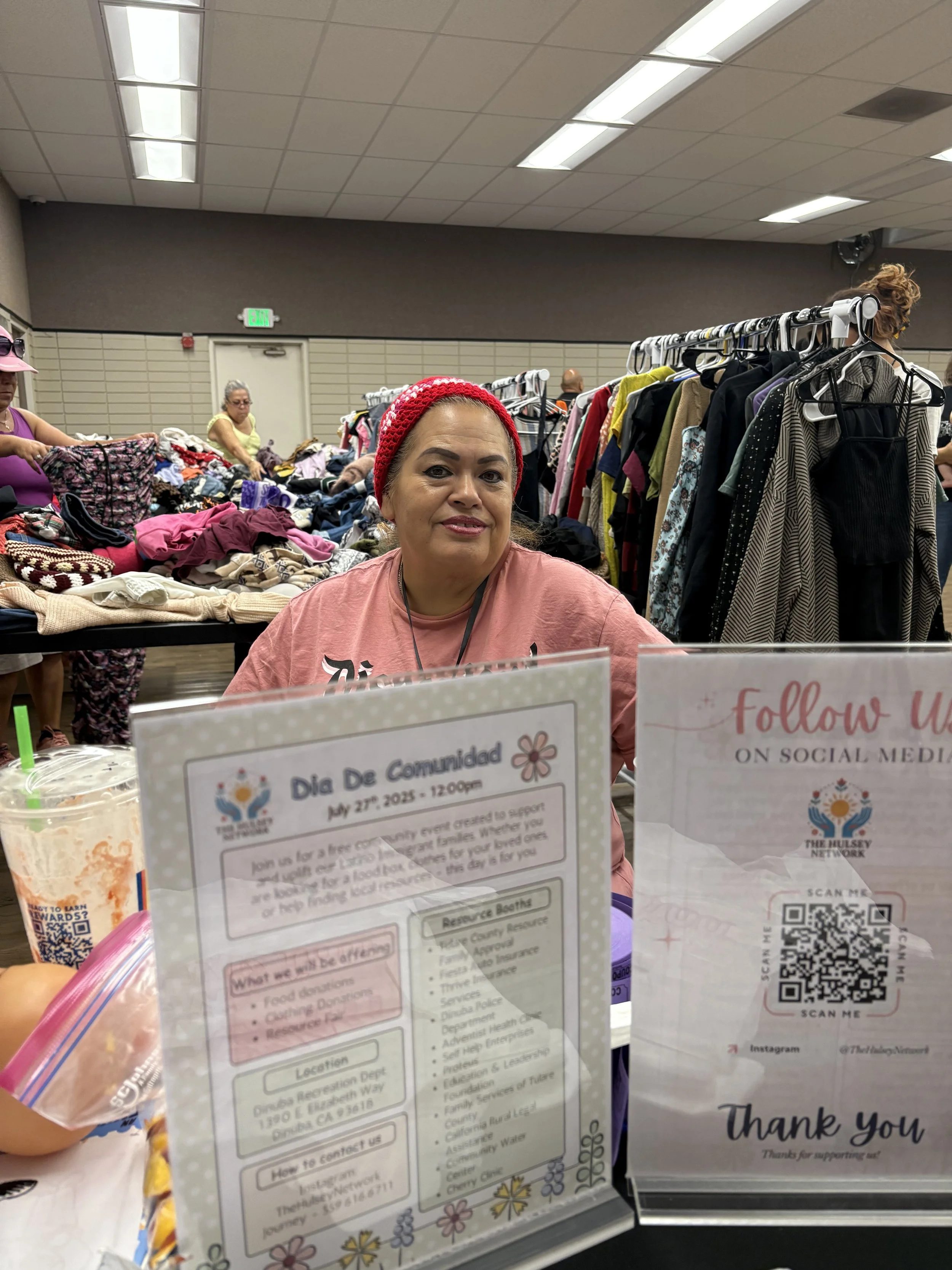 A woman sitting at a table with informational signs about a community event, surrounded by clothing and people shopping in a room with a tiled ceiling.