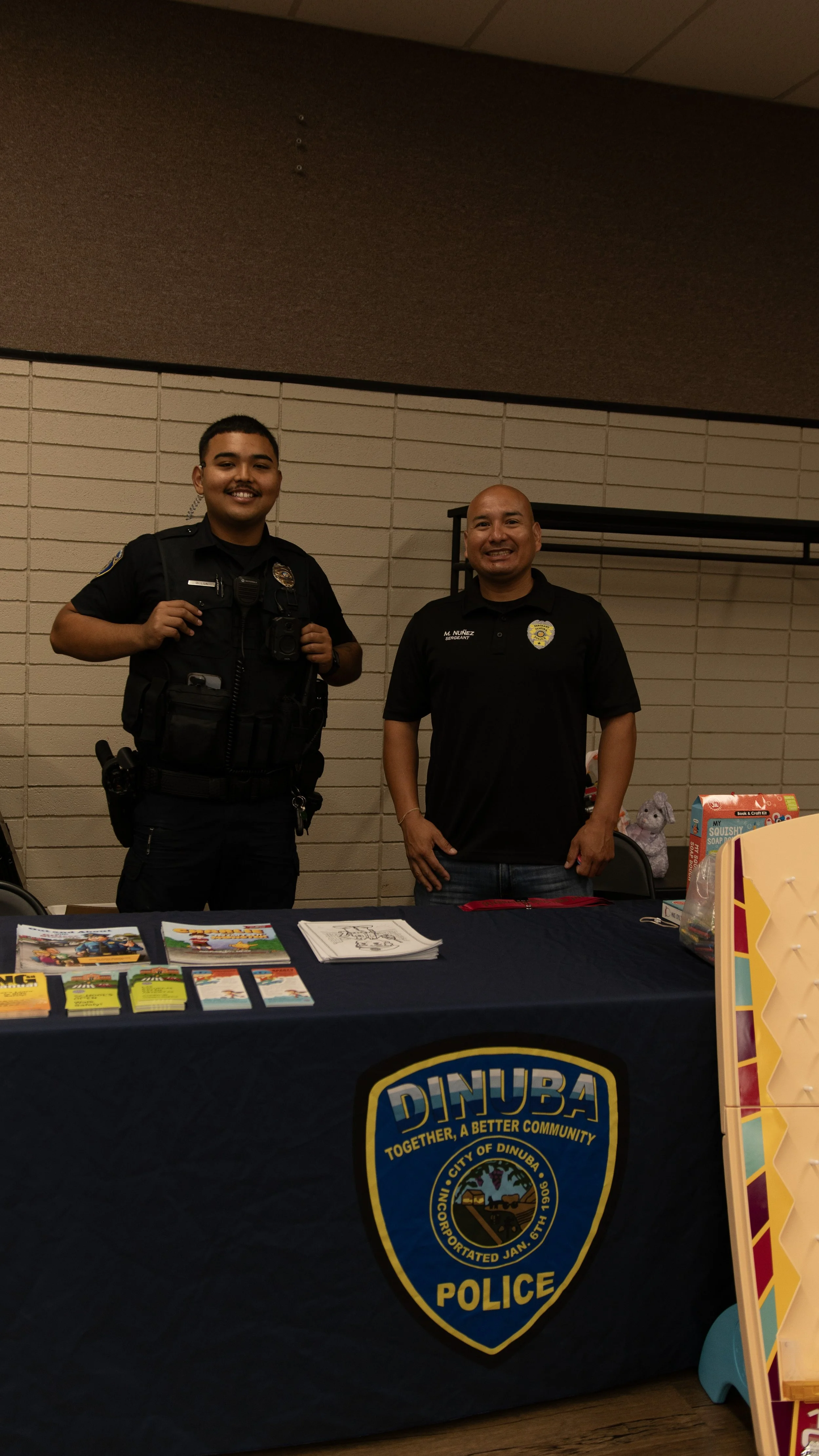 Two police officers standing behind a table with informational pamphlets, flyers, and coloring sheets, representing the Dinuba Police Department at a community event.