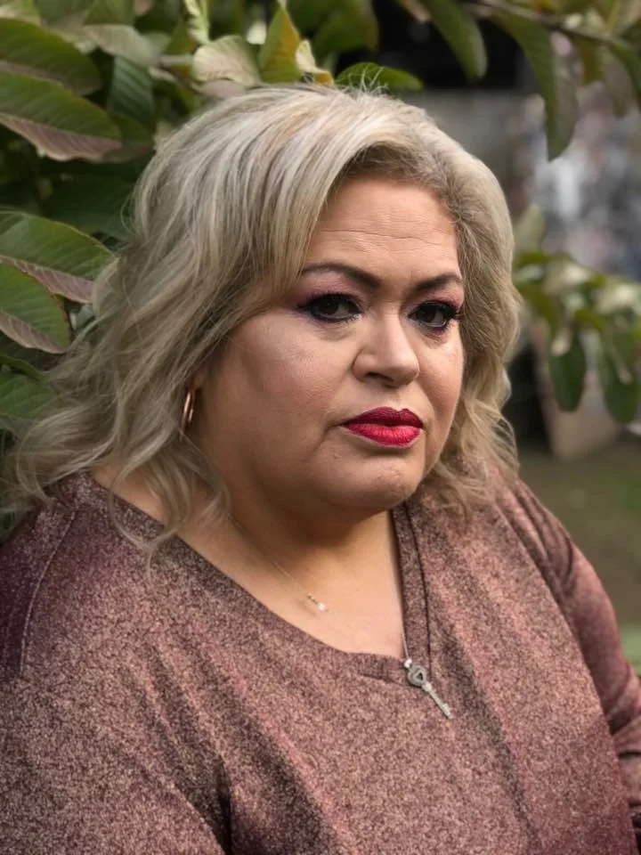 A woman with blonde, wavy hair and red lipstick, wearing a brown top and a necklace with a key pendant, standing outdoors near green foliage.