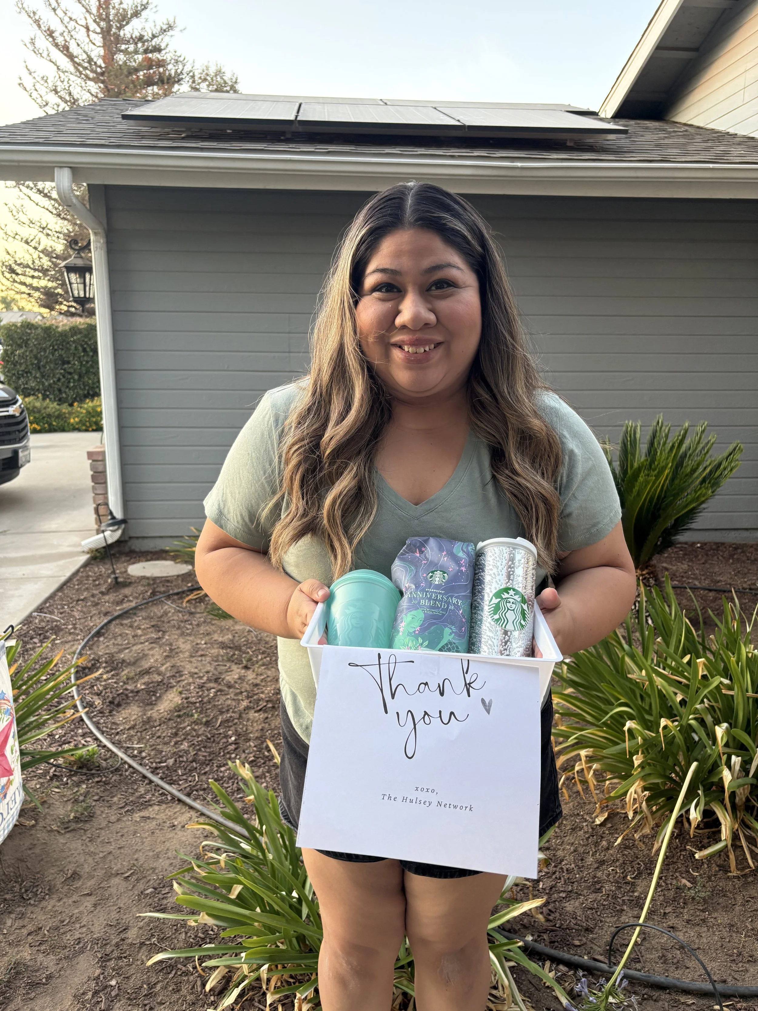 A woman standing outdoors in front of a house holding a white box with Starbucks cups and a box, with a handwritten 'Thank You' card attached. The woman is smiling, has long wavy hair, and is wearing a light green t-shirt and black shorts.