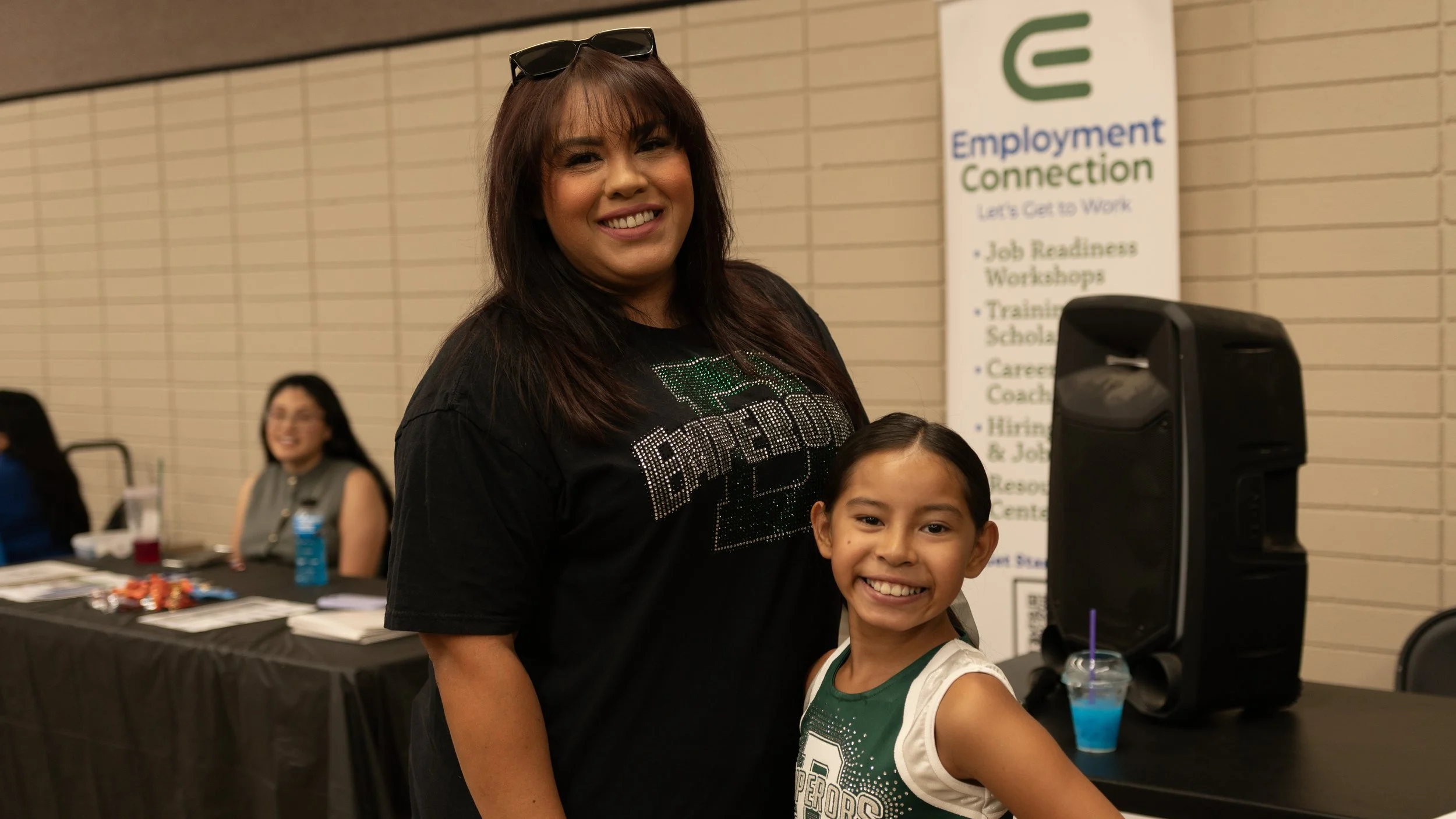 A woman smiling stands next to a young girl, both smiling, at an indoor event with a table and a speaker in the background.
