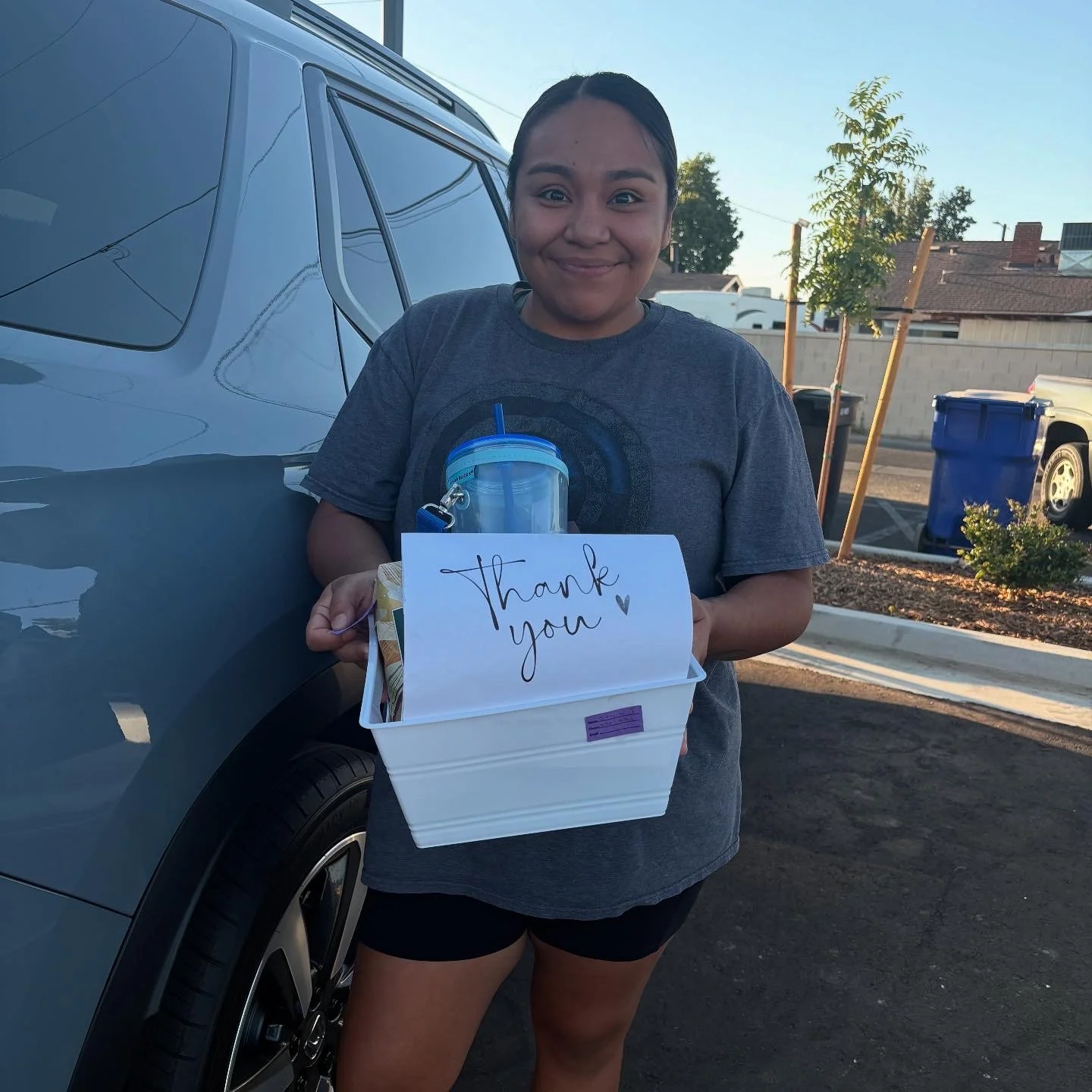 A young woman holding a white box with a 'Thank you' note and a blue tumbler, standing beside a car outdoors during daylight.