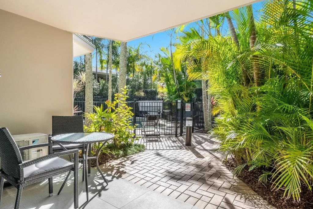 Patio area with outdoor chairs and table, surrounded by lush green tropical plants, with a black gate in the background and a clear blue sky overhead.