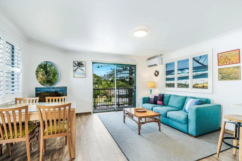 Living room with a blue sofa, wall art of beach scenes, a wooden coffee table, and a dining area with wooden chairs, with a sliding glass door opening to a balcony with outdoor greenery.
