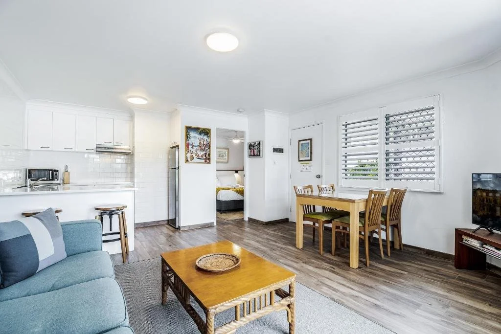 Living room with a light blue sofa, wooden coffee table, and dining table with chairs near two windows with white blinds. Kitchen area in the background with white cabinets and a small refrigerator.