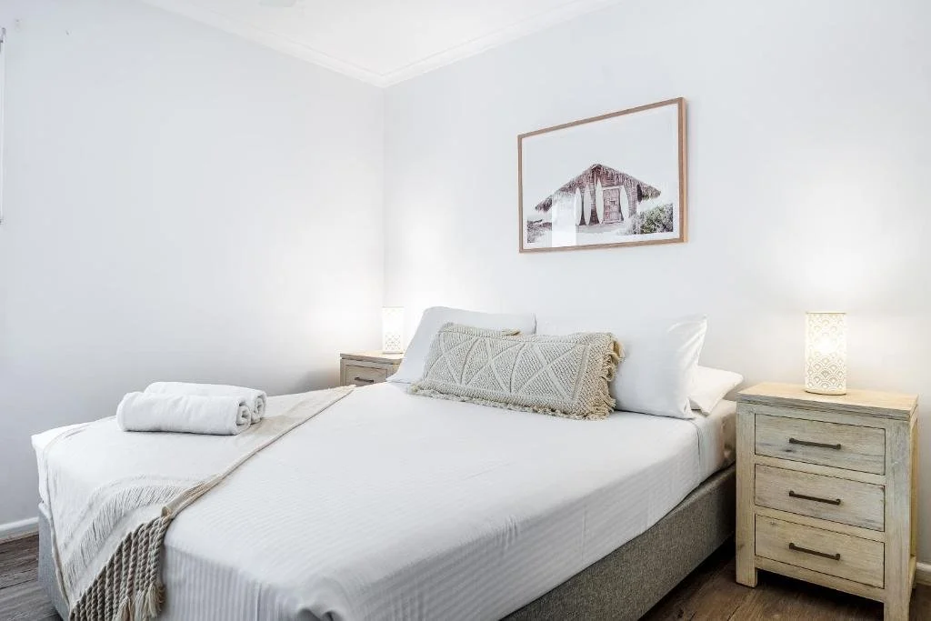 A neatly made bed with white linens, a beige decorative pillow, and a rolled towel on top. Flanking the bed are two nightstands with matching lamps, and above the bed hangs a framed picture of a rustic building.
