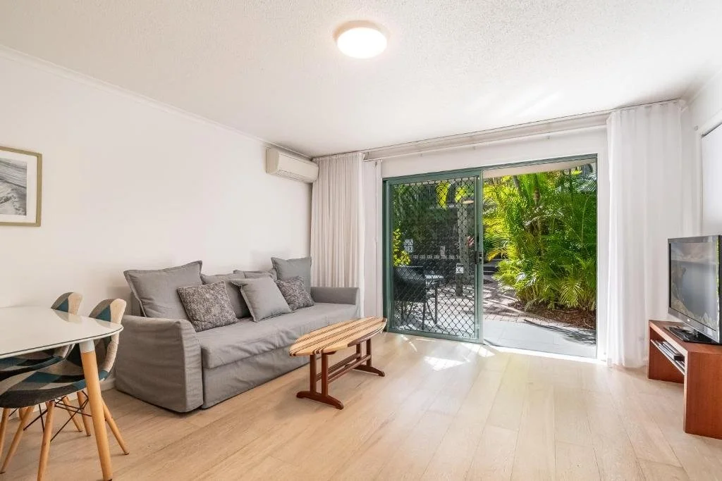 Living room with gray sofa, wooden coffee table, small dining table with chairs, TV on wooden stand, sliding glass door opening to a lush green garden.