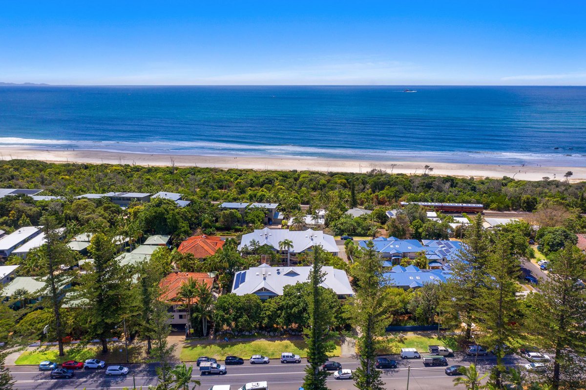 Aerial view of a coastal neighborhood with houses, trees, and parked cars along a street, leading to a sandy beach and the ocean under a clear blue sky.