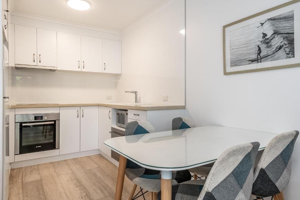 Small kitchen with white cabinets, a built-in oven, microwave, and a white dining table with four chairs, and wall art of surfers.