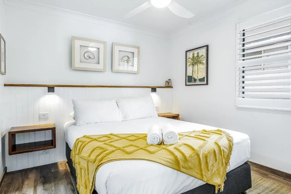 Clean bedroom with white walls, bed with white linens, yellow throw blanket, two towels, wall art, window with white blinds, and wooden nightstands.