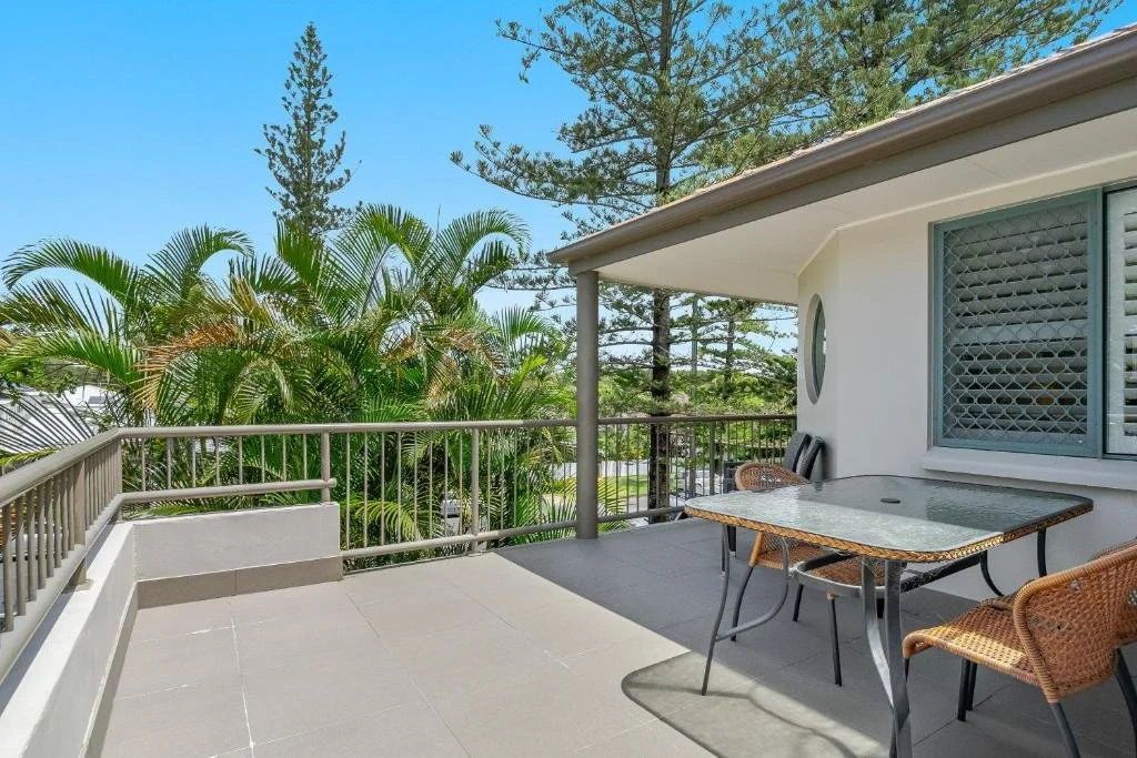 Balcony with glass-top table, wicker chairs, and a view of trees with pine and palm.