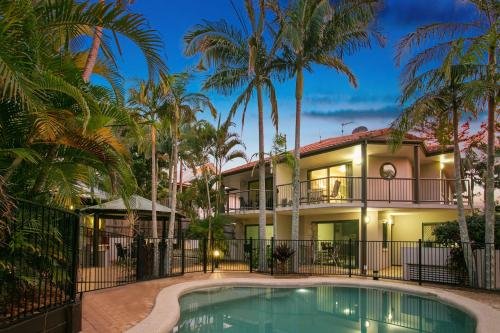 A tropical residential backyard with a swimming pool, surrounded by palm trees and a two-story house with exterior lighting during dusk.