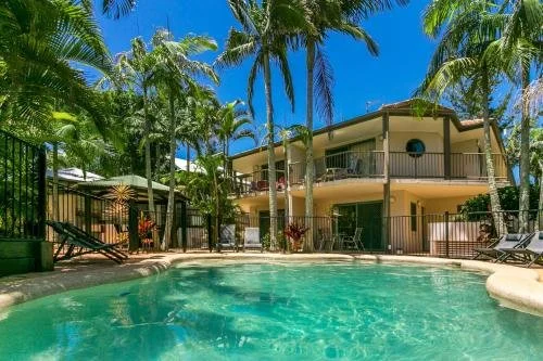 A luxurious backyard with a swimming pool surrounded by palm trees, lush greenery, and a two-story house with a balcony.