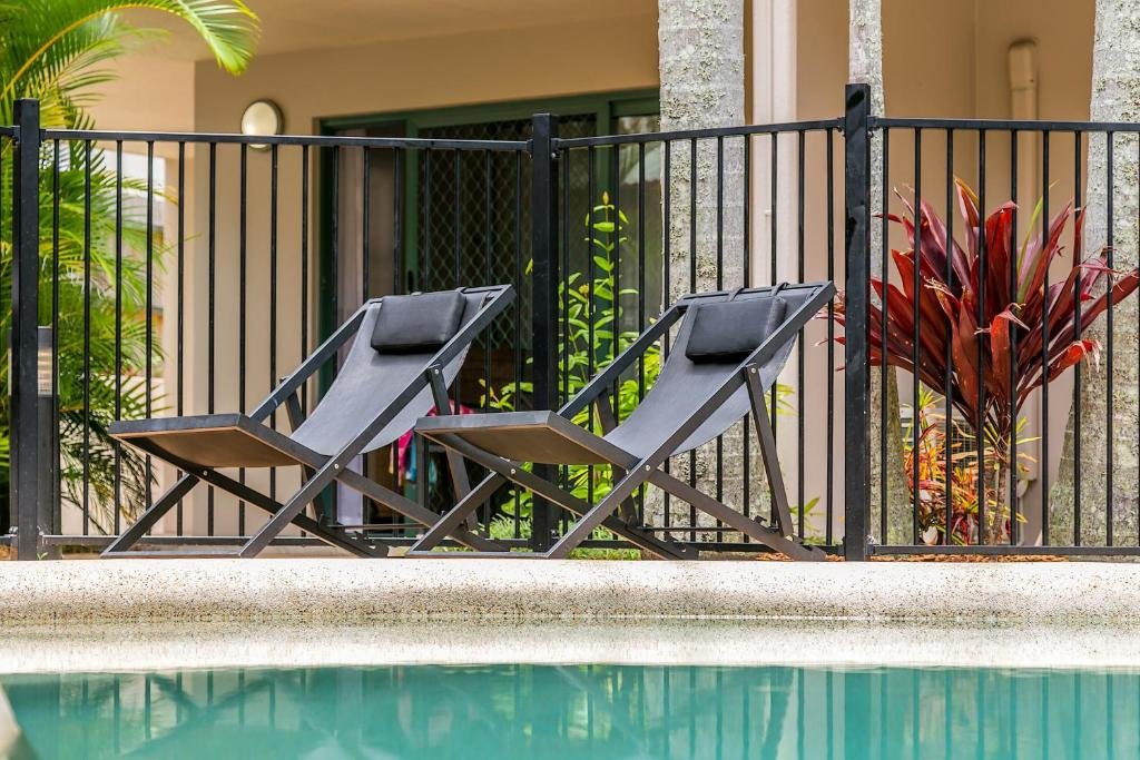 Two black lounge chairs by a swimming pool, positioned in front of a black metal fence with tropical plants in the background.