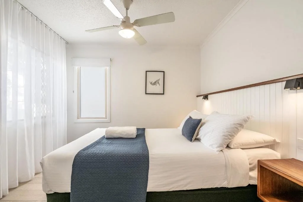 A minimalist bedroom with white walls, a white ceiling fan, and sheer white curtains. There is a bed with white bedding, navy blue pillows, a navy blue quilt, and a wooden nightstand.