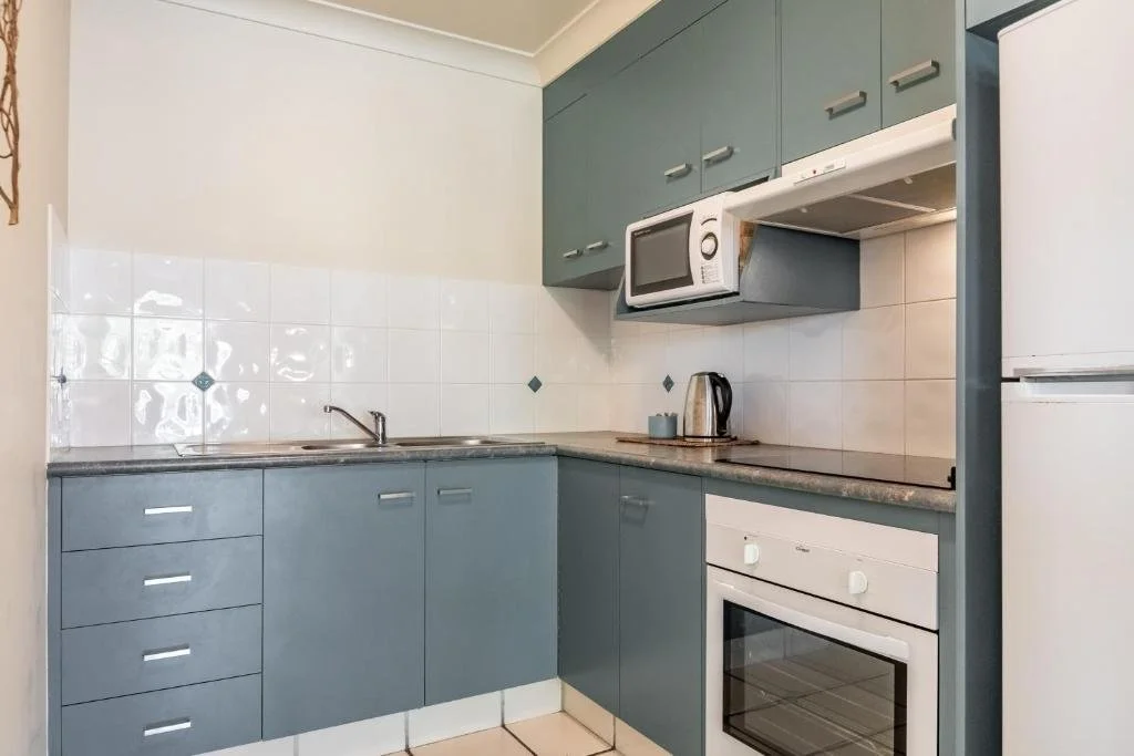 A kitchen with blue cabinets, white tile backsplash, and white appliances including a microwave, oven, and refrigerator. There is a small counter with a kettle and mug.