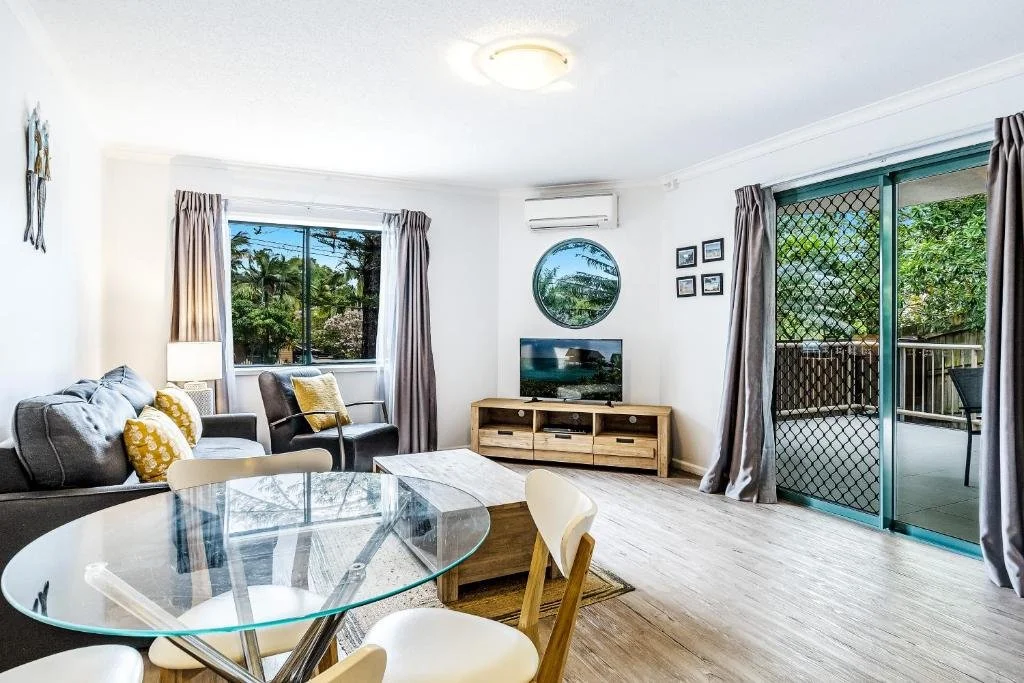 Bright living room with gray sofas and yellow cushions, glass dining table, wooden TV stand with TV, large windows with curtains, sliding glass door to outdoor balcony, and a circular mirror on the wall.