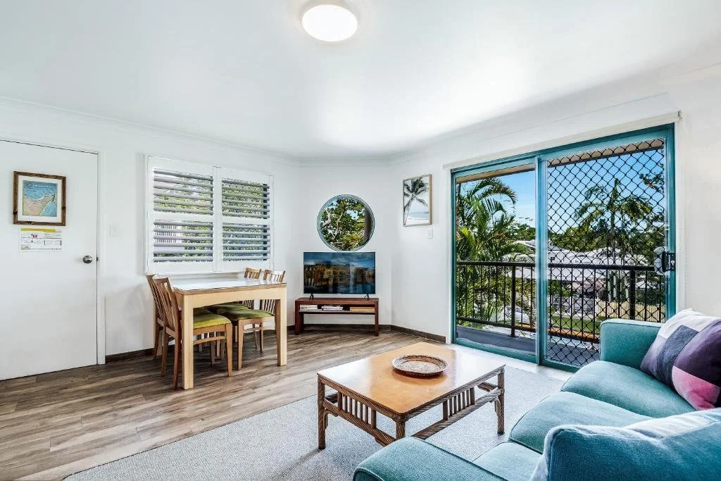 Living room with a teal sofa, wooden coffee table, and a TV on a stand. There is a dining table with four chairs near a window, and a sliding glass door leading to a balcony with a view of trees.