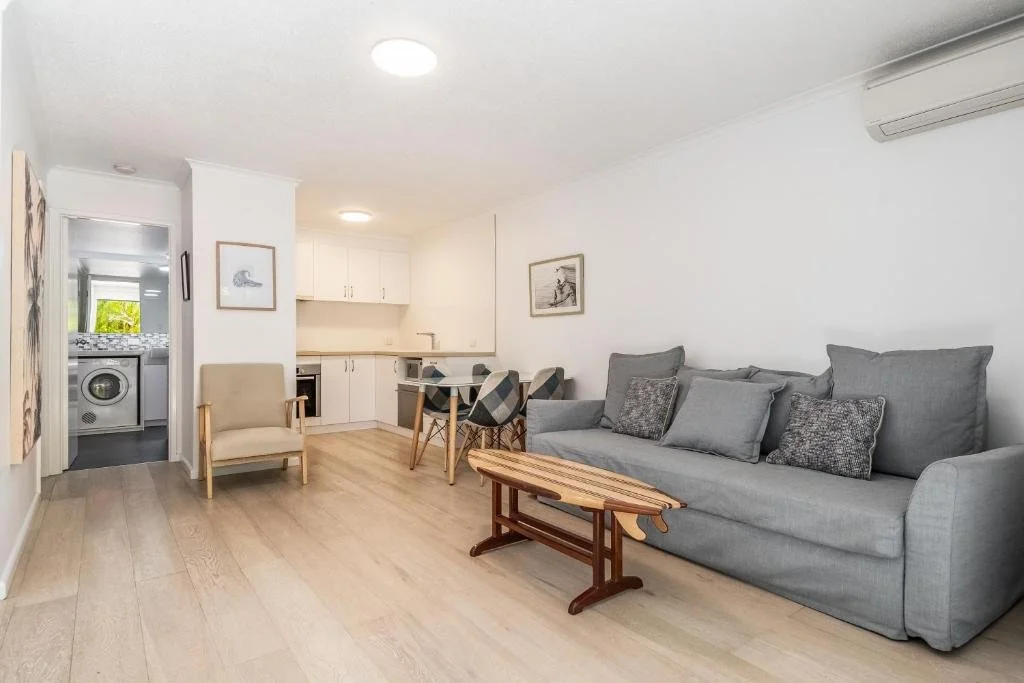 Living room with gray sofa, beige armchair, small wooden table, and dining area with chairs, kitchen in the background, white walls, light wood flooring, and framed artwork.