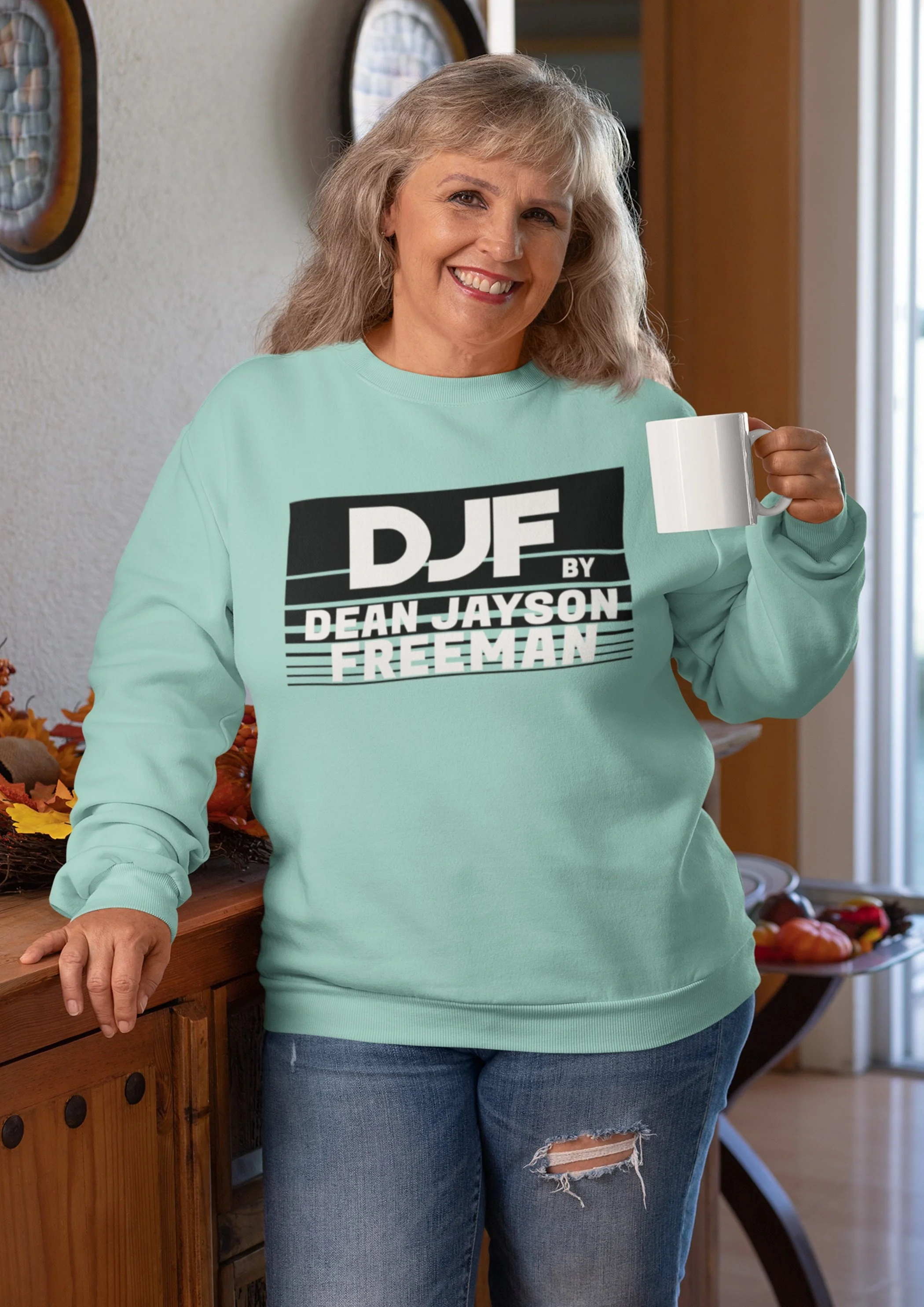 Ein lächelnde Frau in einer hellgrünen Sweatshirt mit schwarzen und weißen Text, die in einer Küche steht, während sie eine weiße Tasse hält. Sie trägt Jeans mit einem Riss im Knie.