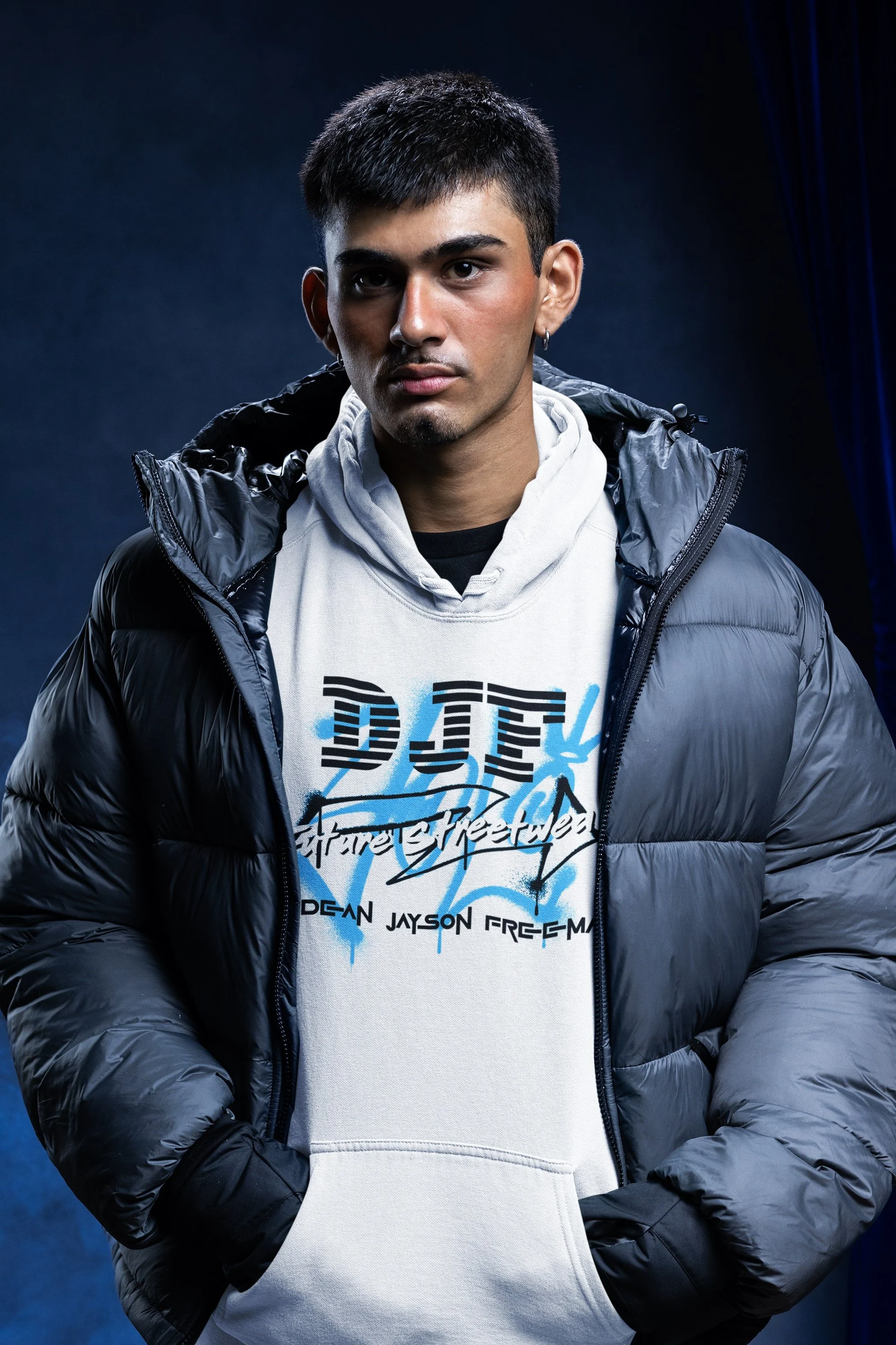 Junge Mann trägt eine schwarze Winterjacke über einem weißen Hoodie mit graffitiähnlichem Design und Text, vor dunklem Hintergrund.