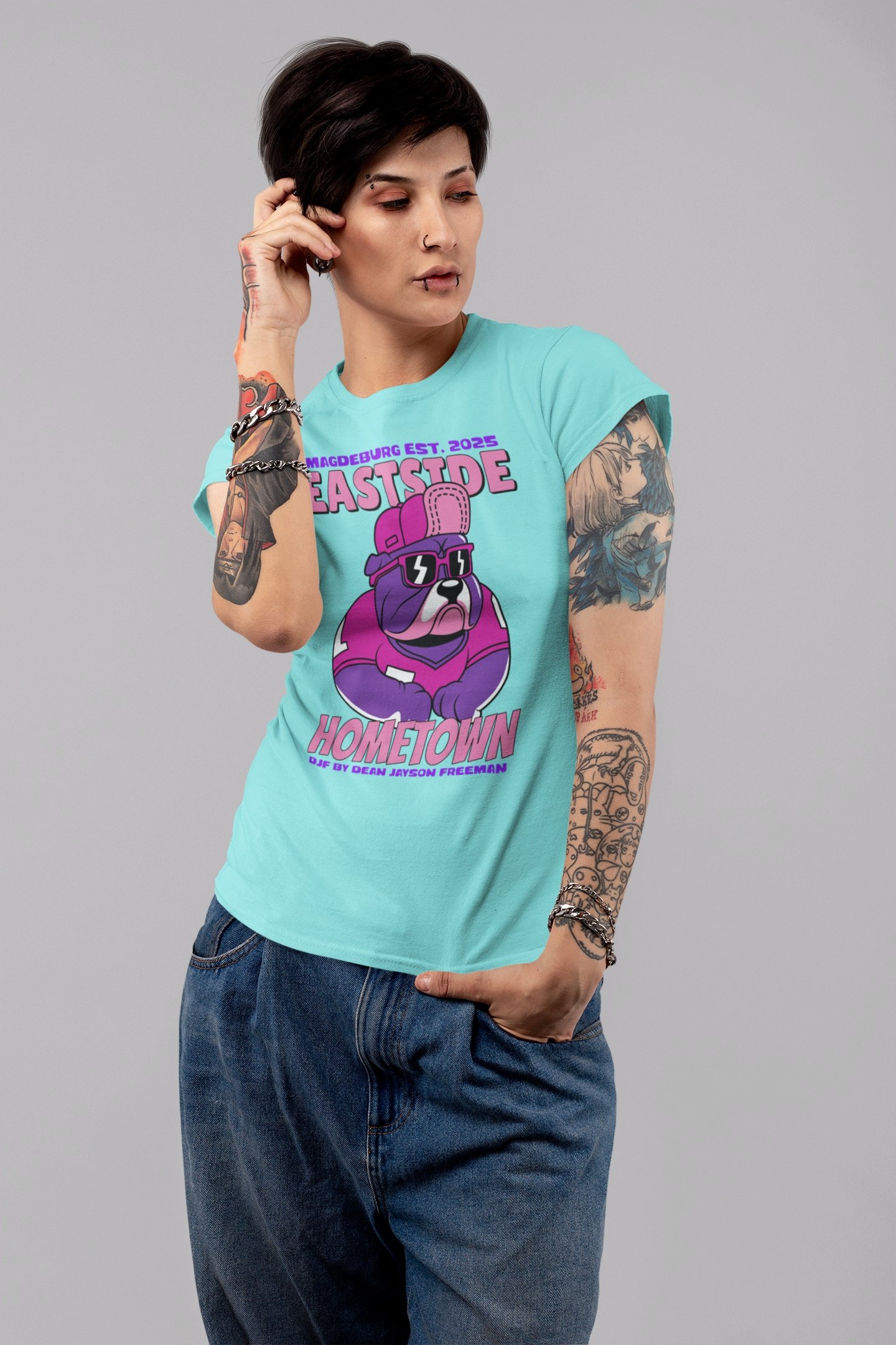 Junge Frau mit kurzen dunklen Haaren, Tattoos an Armen, trägt hellblaues T-Shirt mit Pink und Lila Grafik und Text, Jeans, gestenreiche Pose, neutraler grauer Hintergrund.
