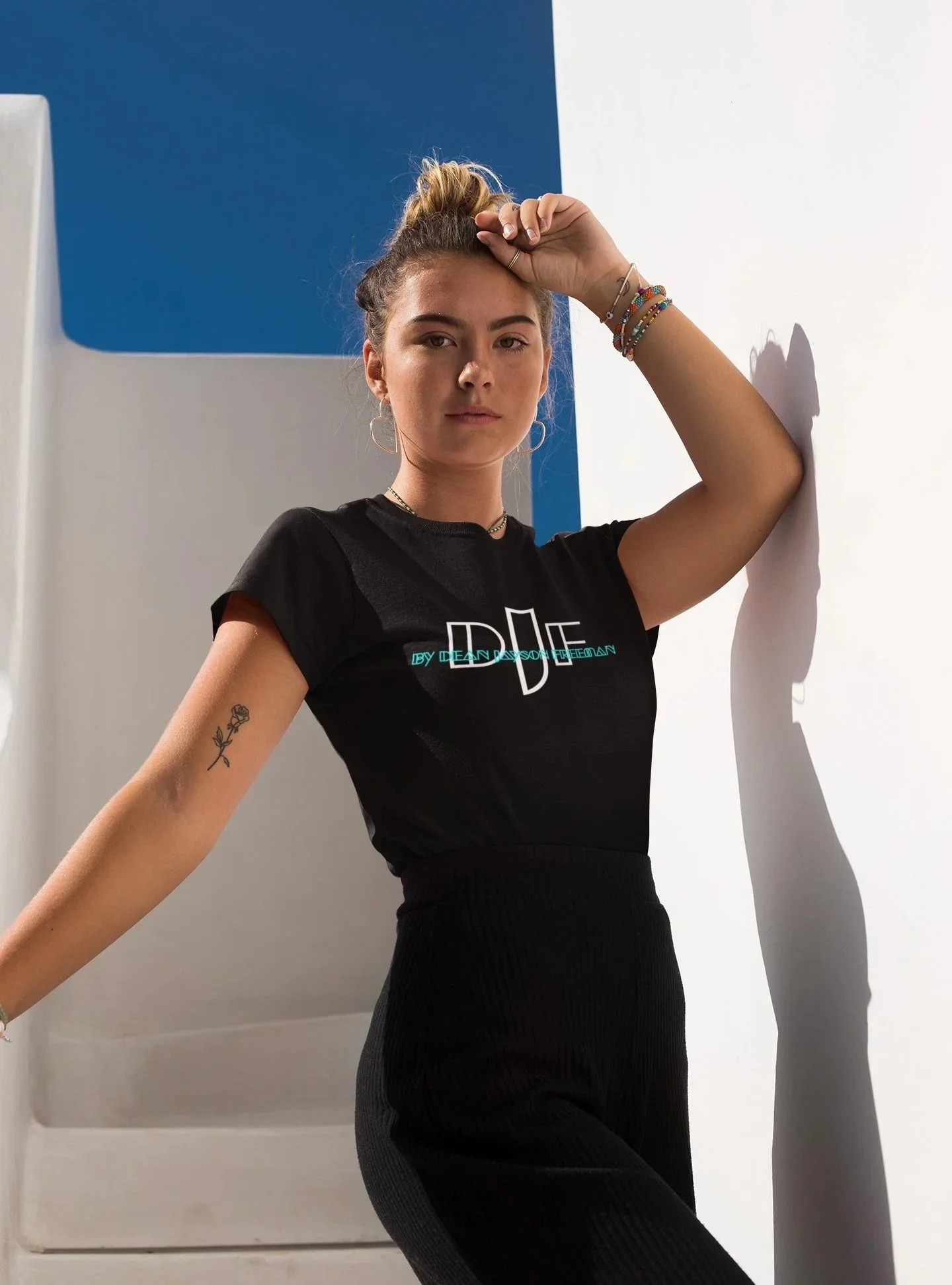 Junge Frau mit Tattoos, Armbändern und dunkler Kleidung lehnt an einer weißen Wand und blickt in die Kamera.